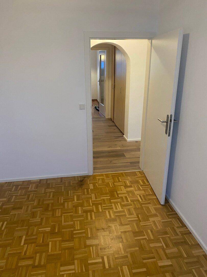 Helles, leeres Zimmer mit Parkettboden und weiß gestrichenen Wänden, das zu einem Flur führt.