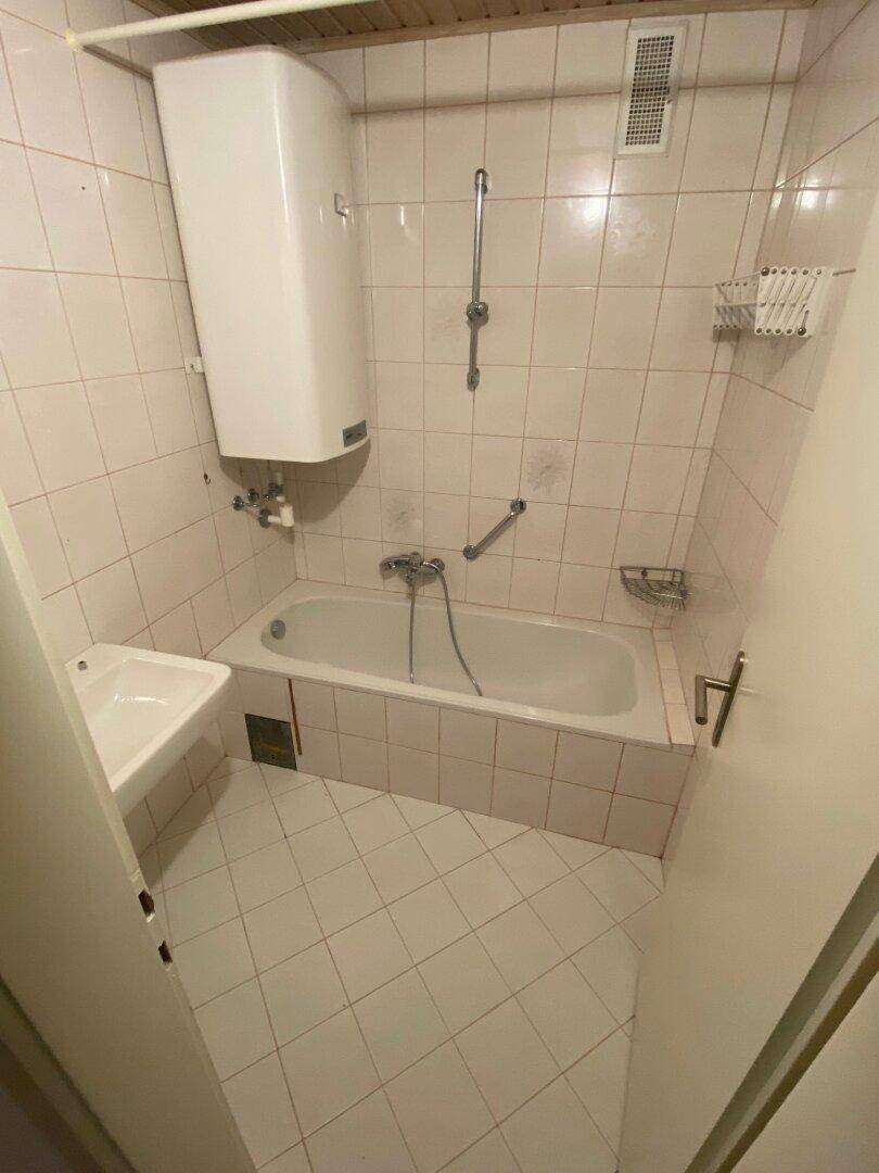 Badezimmer mit weißen Fliesen, einer Badewanne und einem kleinen Waschbecken, ausgestattet mit einem Warmwasserboiler.
