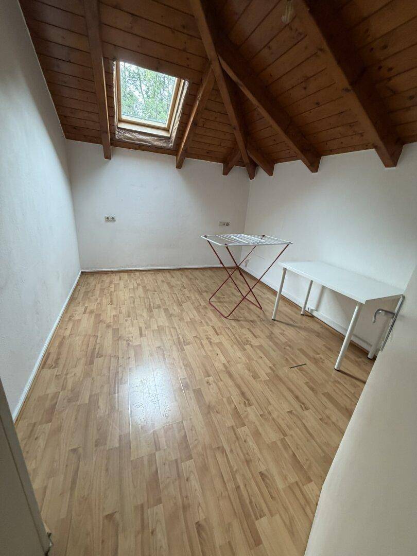 Helles Dachzimmer mit sichtbaren Holzbalken, Dachfenster und Laminatboden.