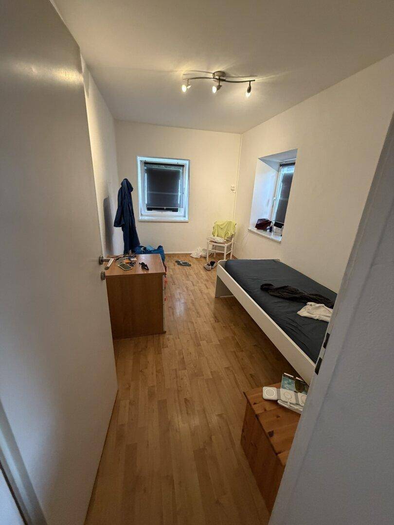 Einfaches Schlafzimmer mit Holzboden und zwei Fenstern, funktional eingerichtet.