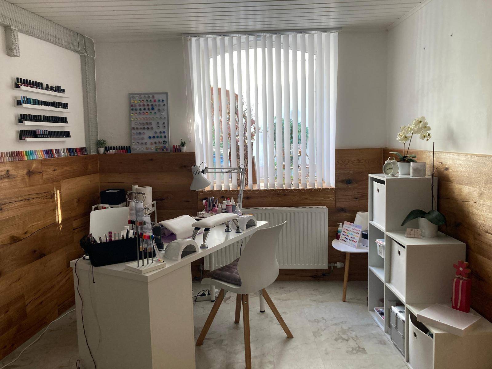 Nagelstudio-Arbeitsplatz mit Fenster, Schreibtisch und Regalen für Nagellacke, hell und funktional.