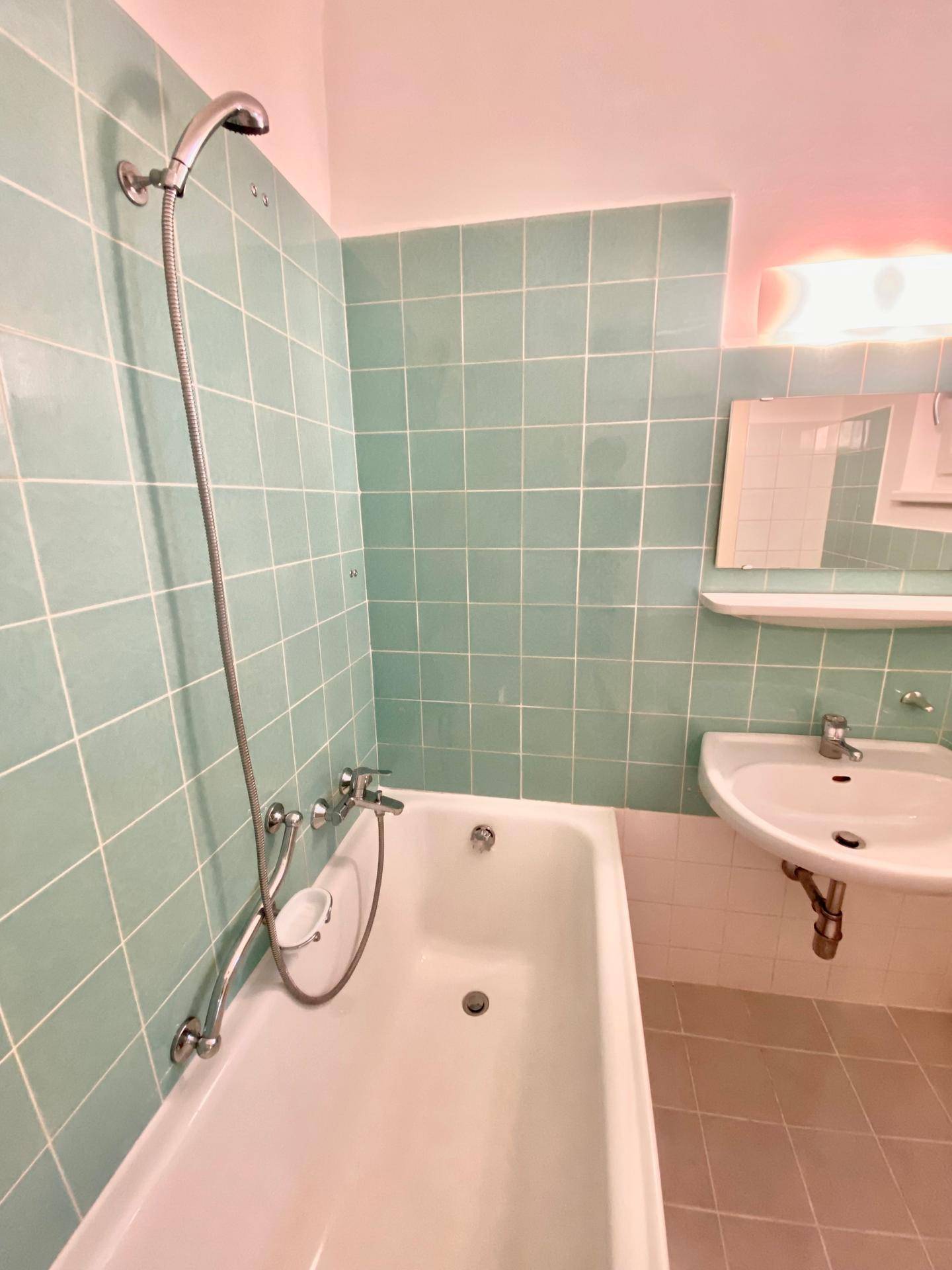 Badezimmer mit hellgrünen Fliesen, Badewanne mit Duschfunktion und Waschbecken.