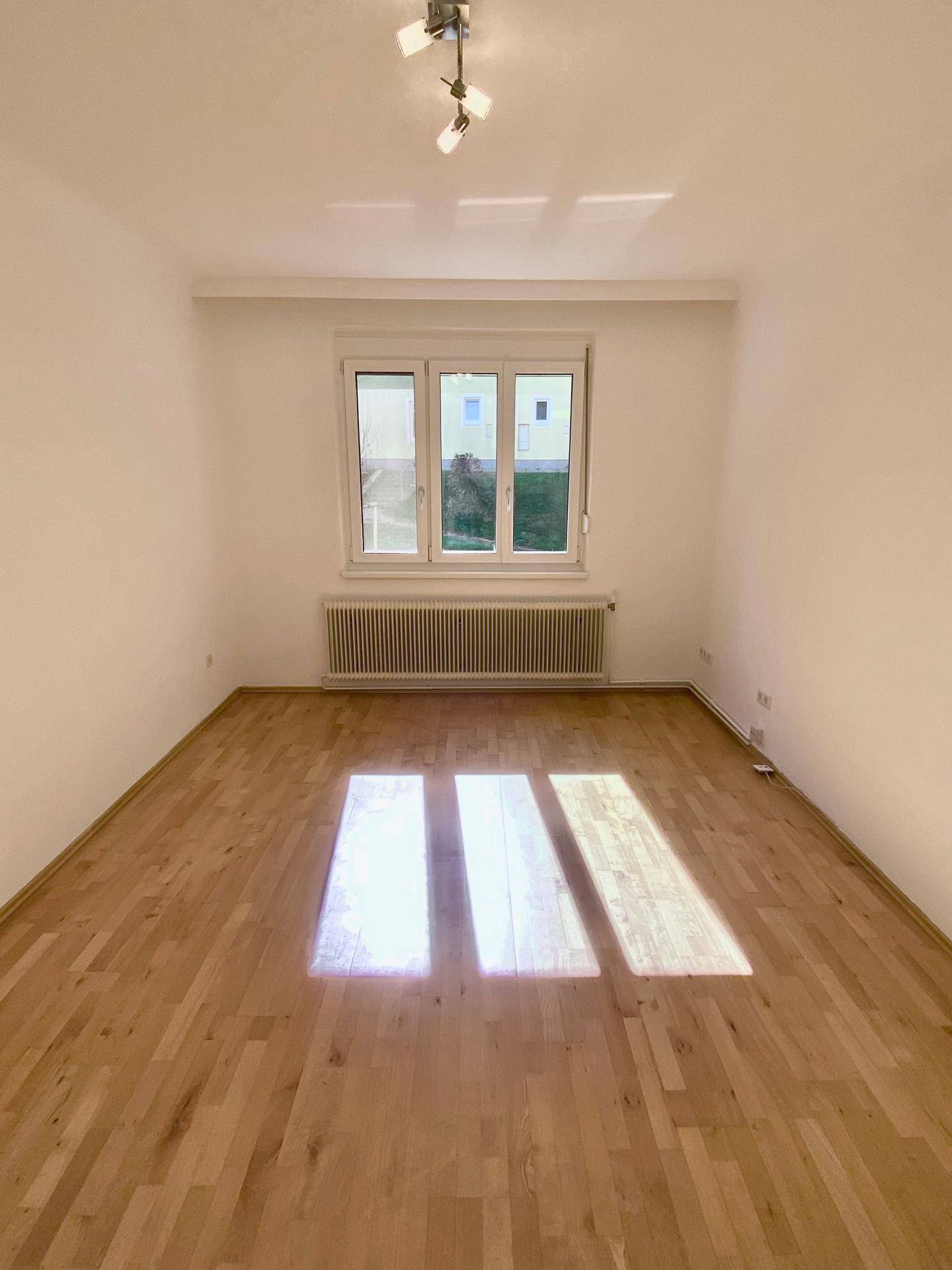 Helles, leeres Zimmer mit Parkettboden und großem Fenster, das viel Tageslicht hereinlässt.