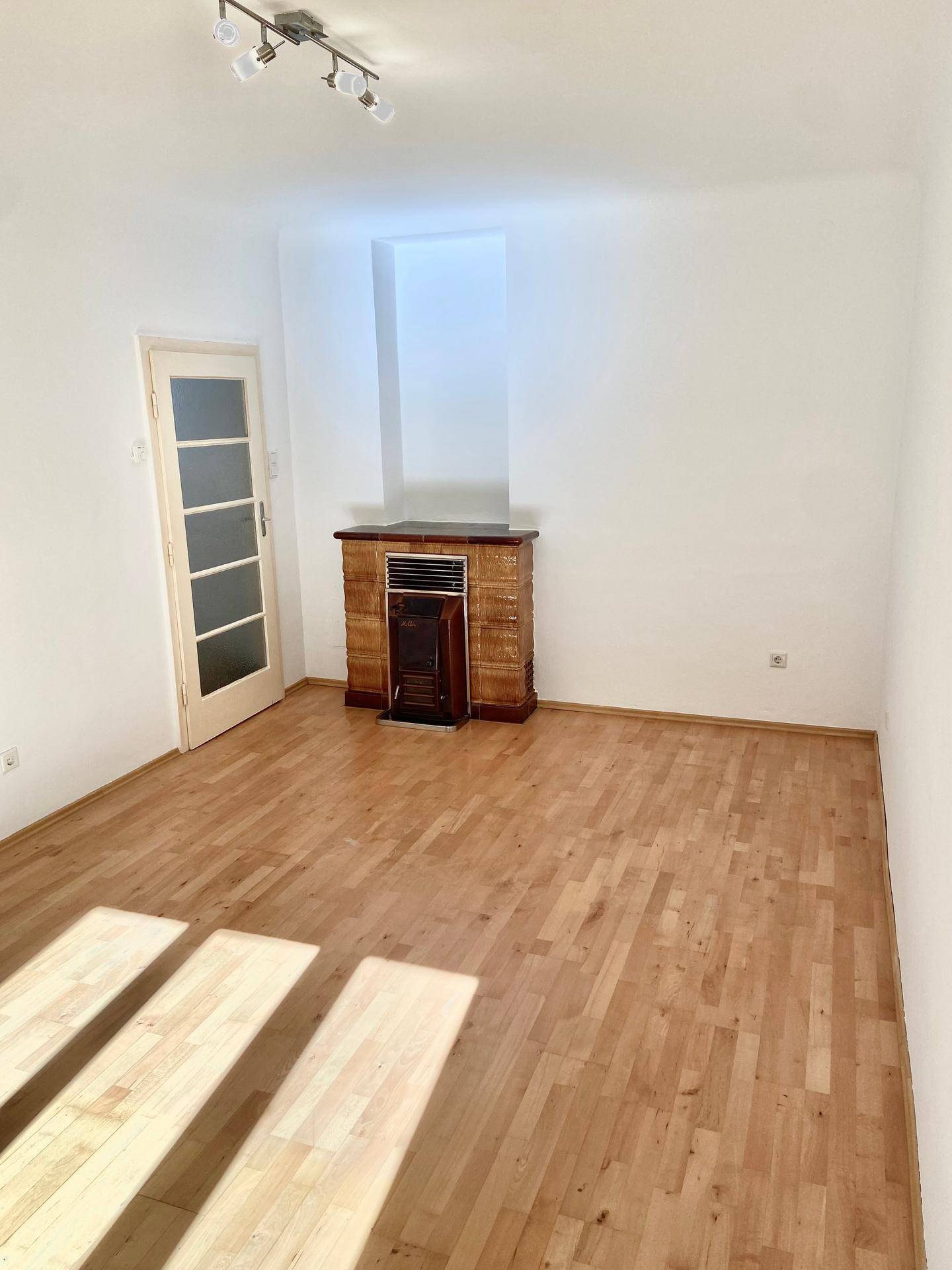 Leeres Wohnzimmer mit hellem Parkettboden und einem traditionellen Kachelofen.