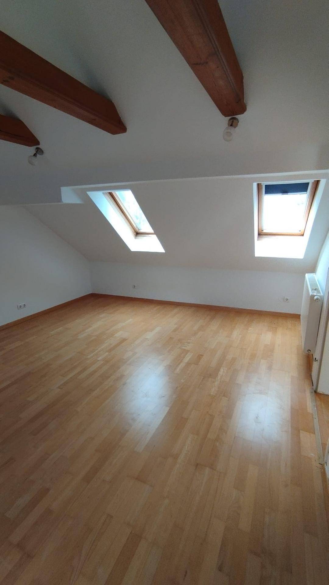 Geräumiges Dachzimmer mit sichtbaren Holzbalken, Parkettboden und zwei Dachfenstern.