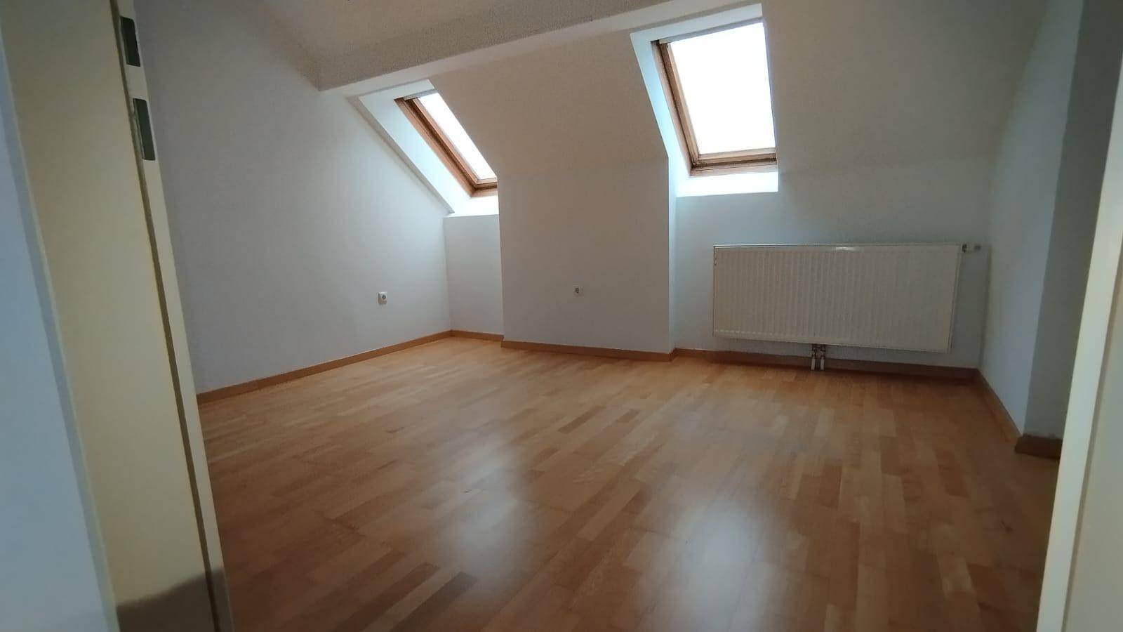 Helles Dachzimmer mit Holzparkettboden, Heizkörper und zwei großen Dachfenstern.