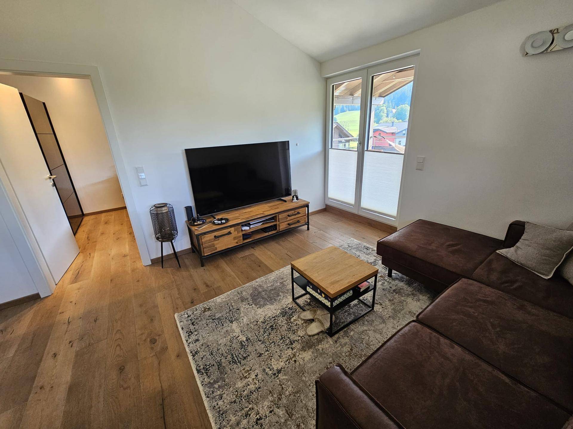 Geräumiges Wohnzimmer mit großem Fernseher, Holzboden und Zugang zu weiteren Räumen.