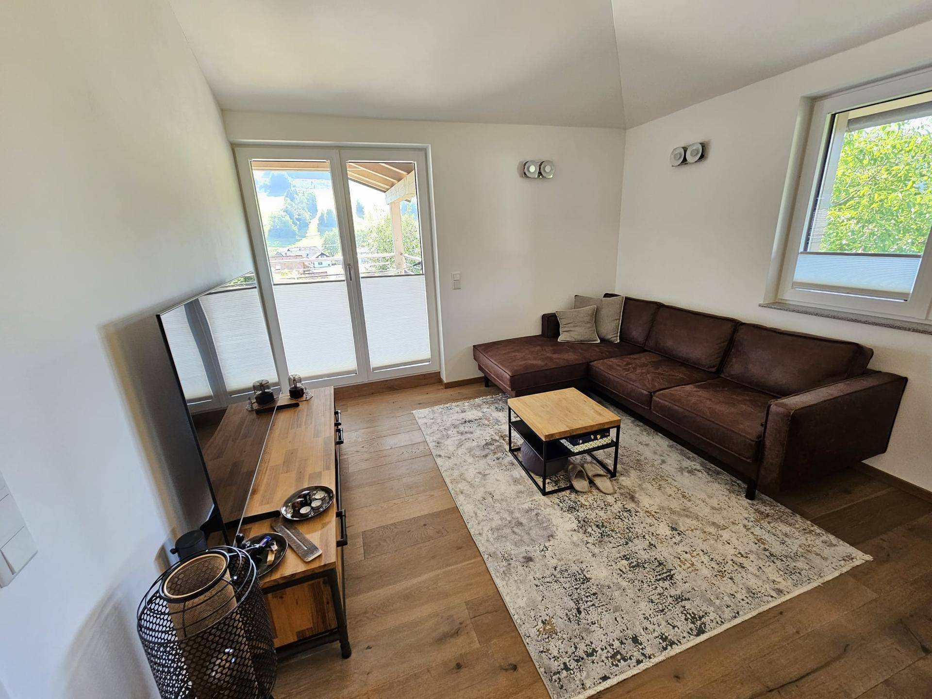 Helles Wohnzimmer mit braunem Ecksofa, Holzboden und Zugang zum Balkon.
