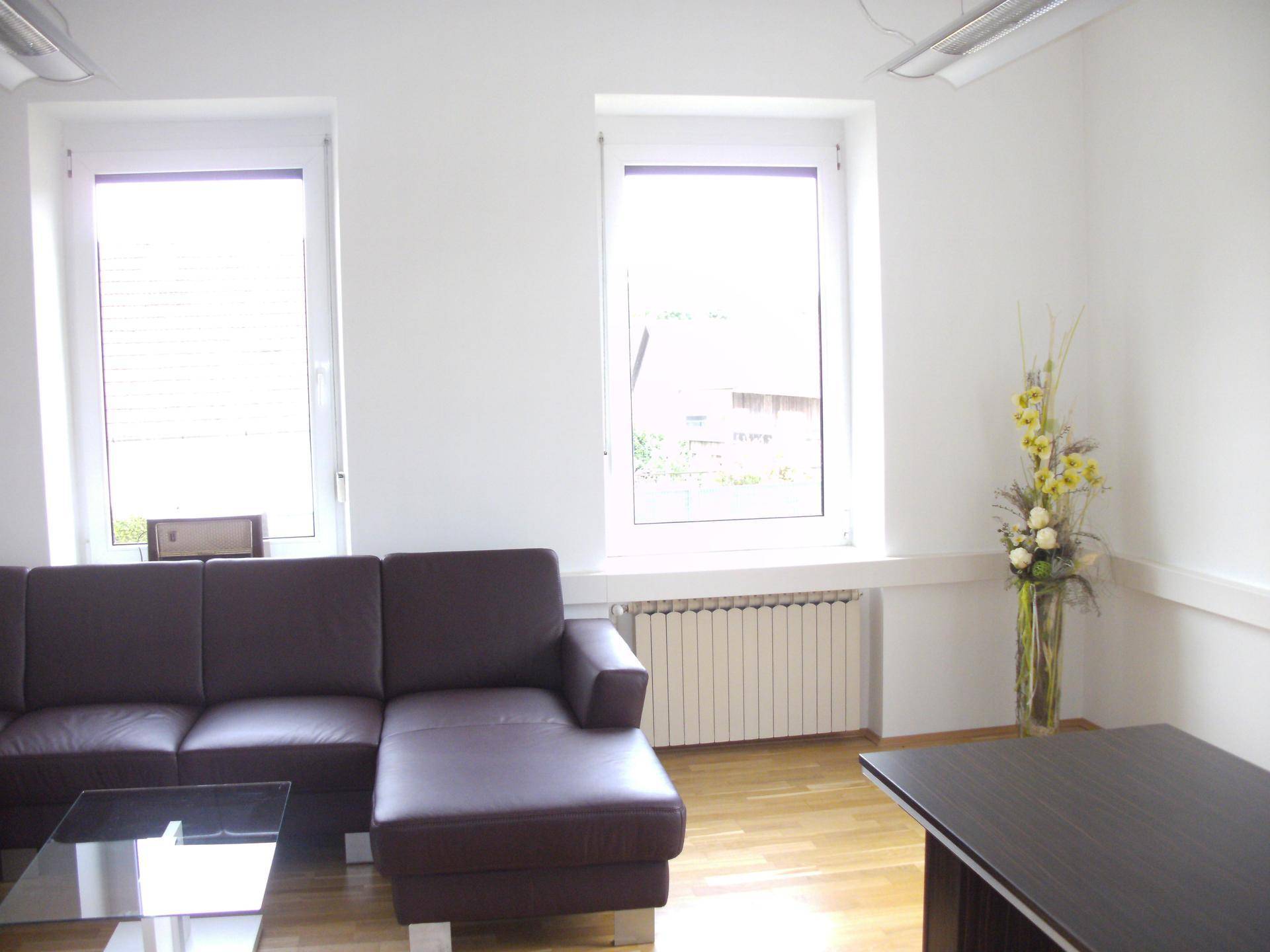 Detailansicht eines Wartebereichs mit Ledersofa, Couchtisch und Heizkörper unter zwei großen Fenstern.