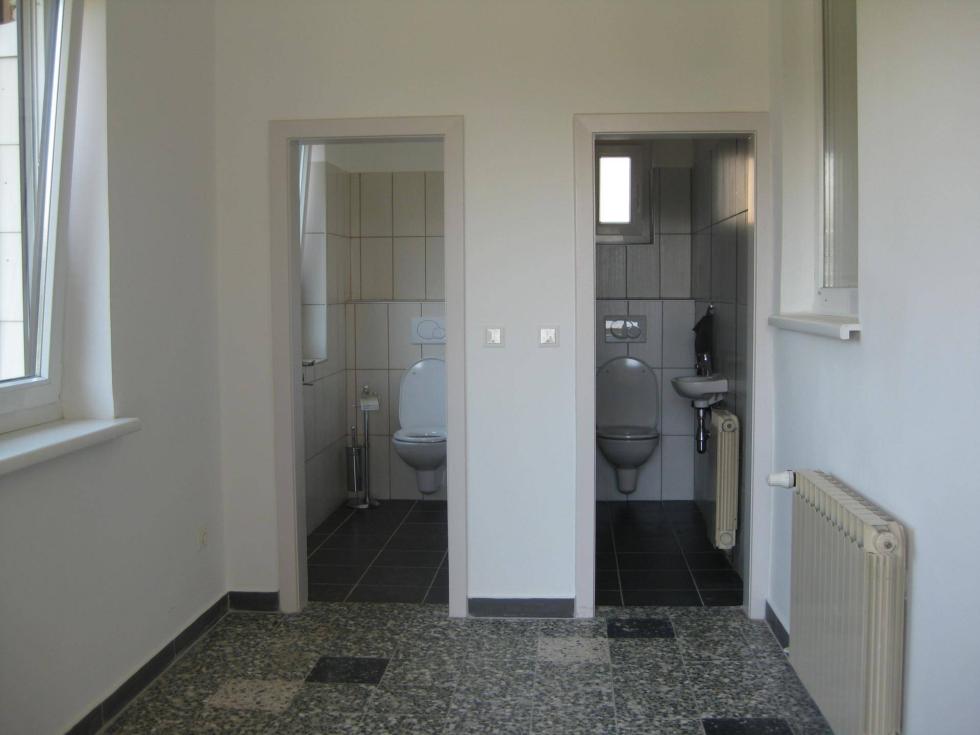 Zwei separate Toilettenräume mit Fliesenboden, weißen Wandfliesen und kleinen Fenstern.