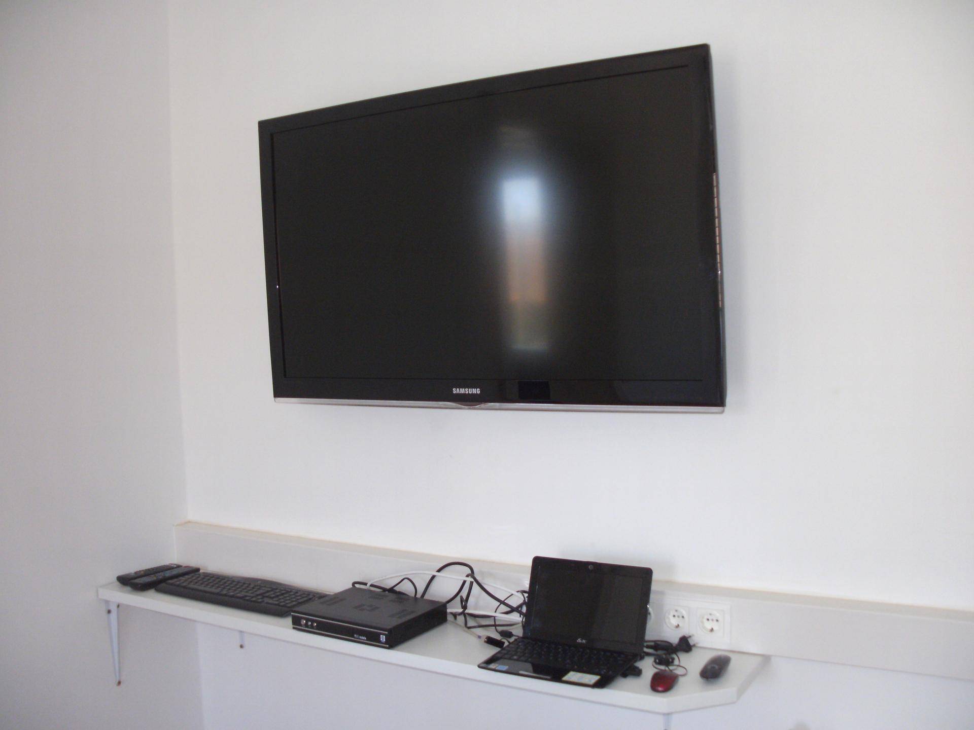 Wandmontierter Flachbildfernseher über einem Regal mit Elektronik und Kabeln in einem hellen Raum.