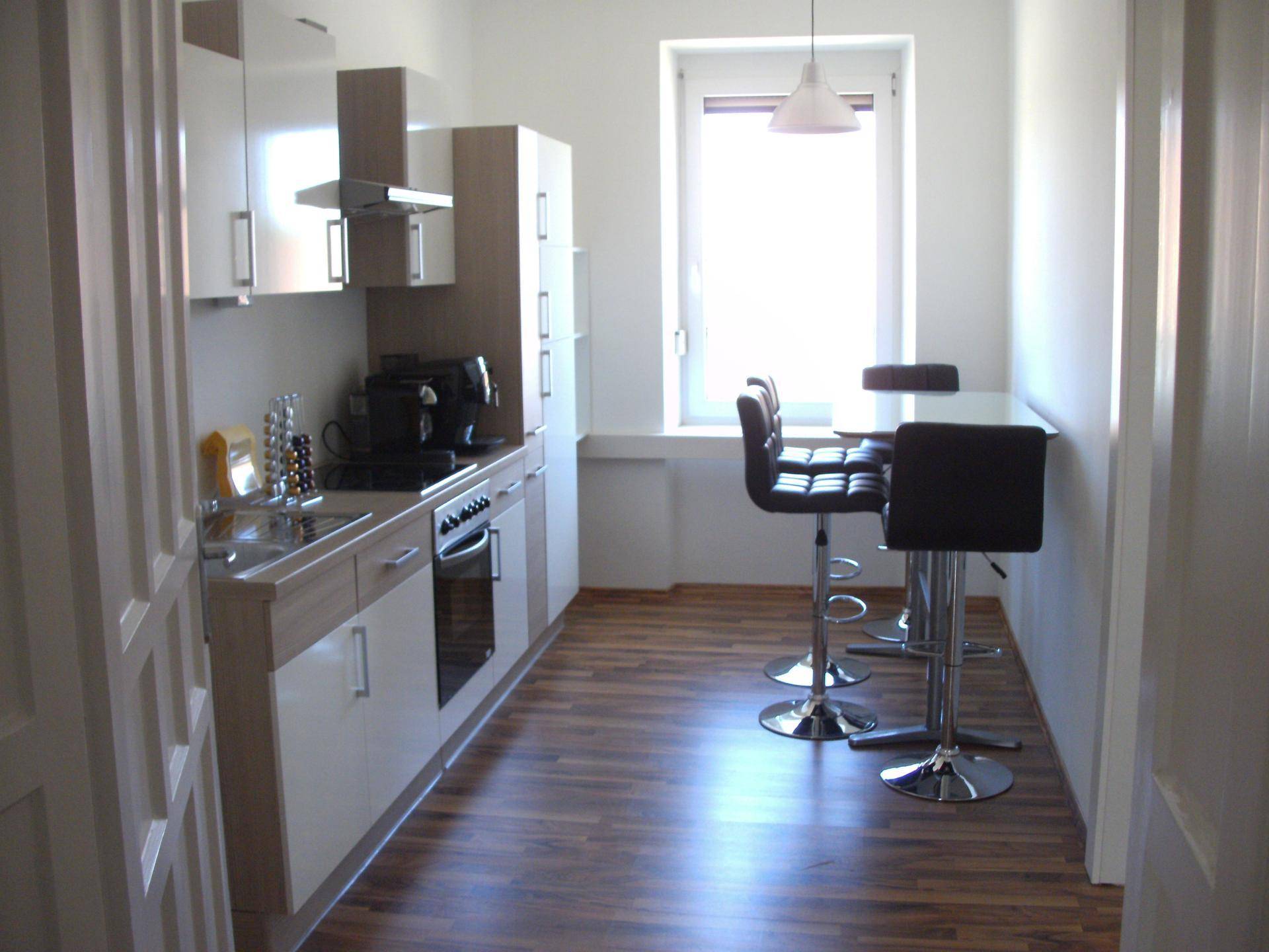 Kleine, moderne Küche mit hellen Schränken, Kochfeld, Spüle und Barhockern an einem Tresen.