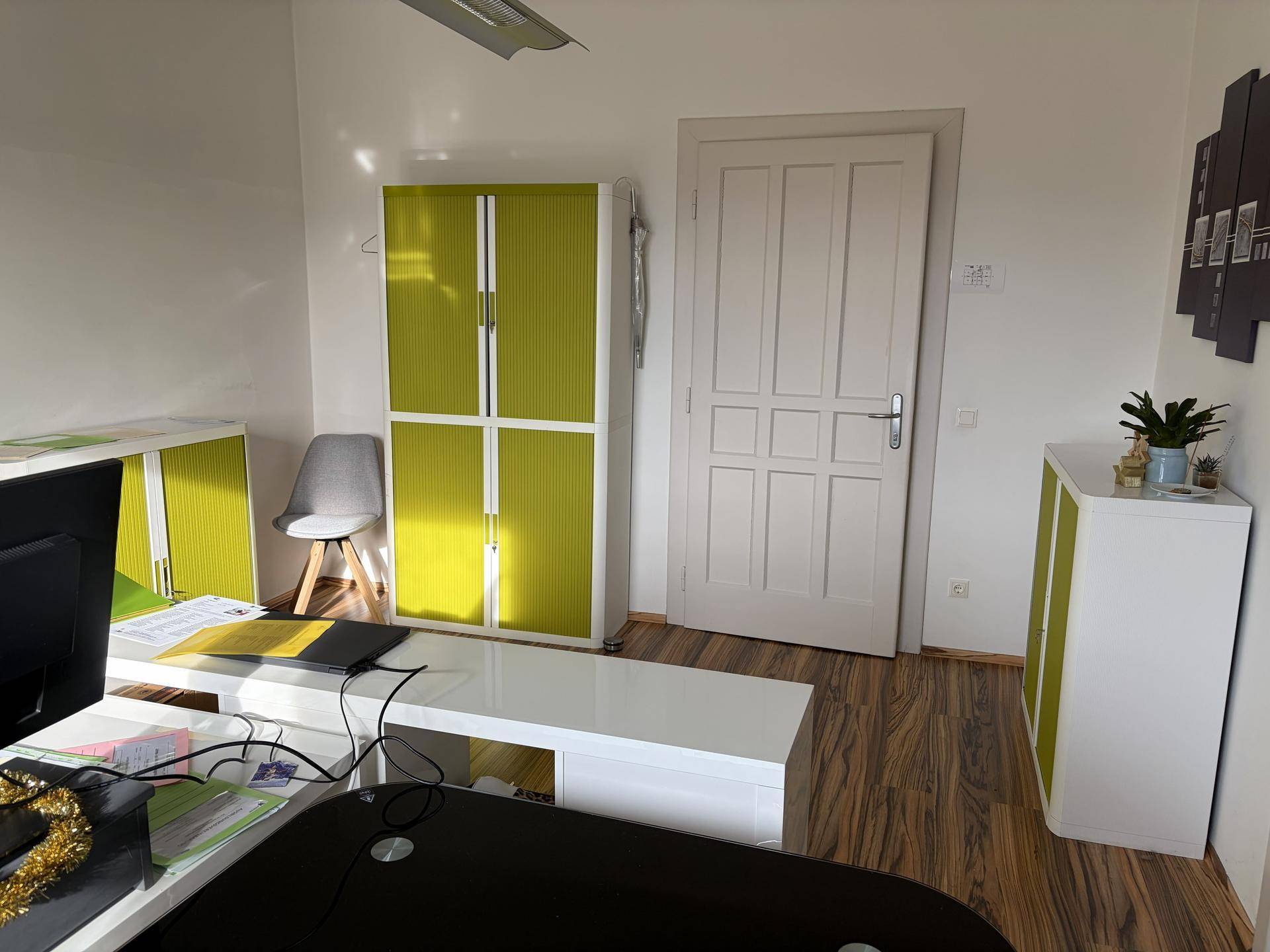 Büro mit weißen Wänden, grün-weißen Schränken und einem Holzlaminatboden, gut beleuchtet.