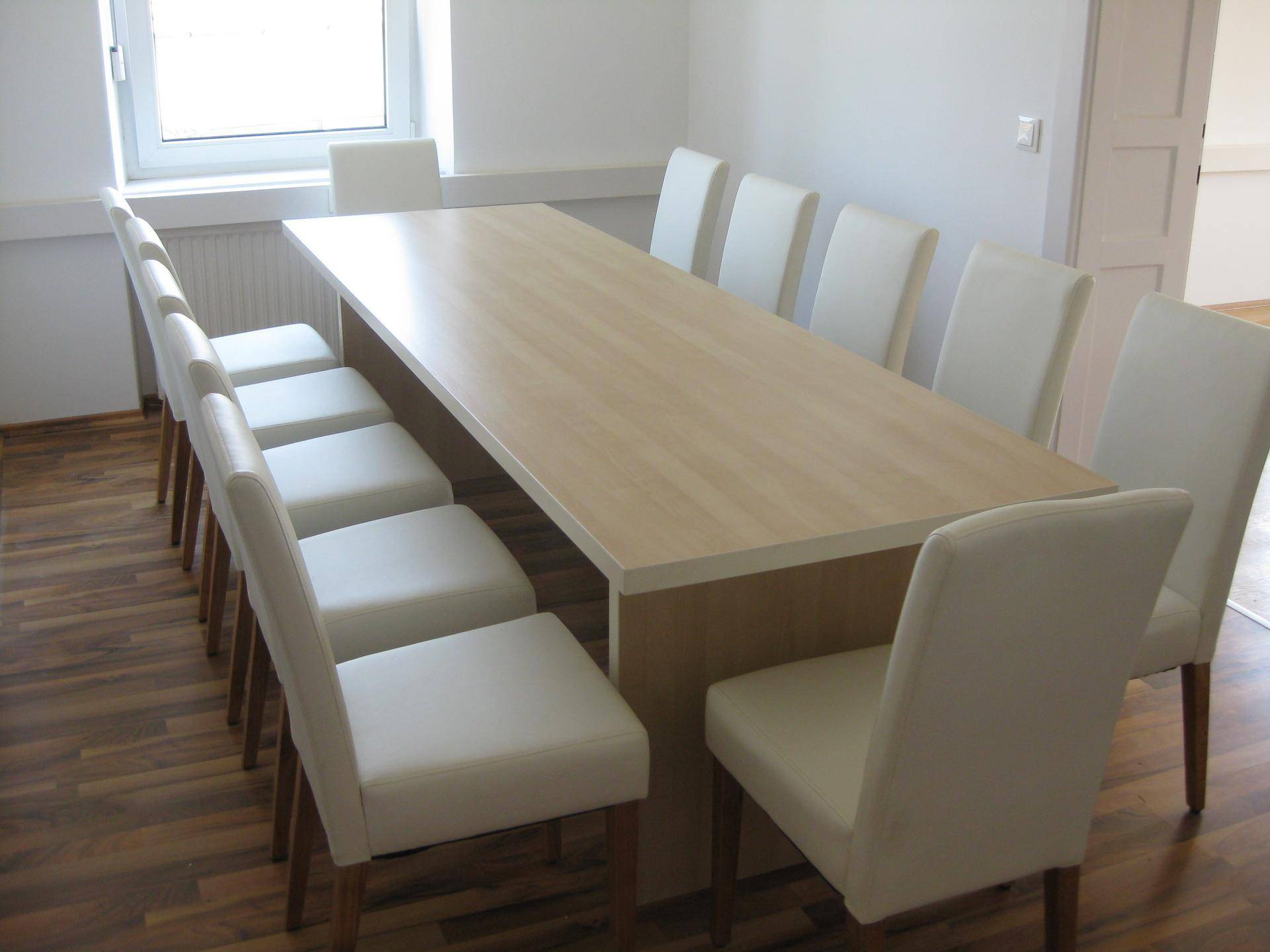 Großer Konferenztisch mit vielen weißen Stühlen in einem hellen Raum mit Holzlaminatboden.