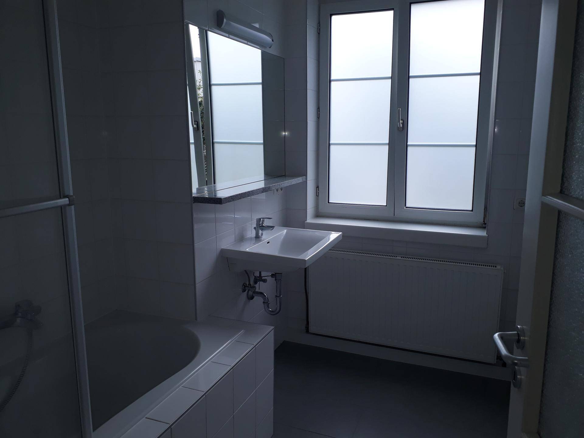 Badezimmer mit weißen Fliesen, Badewanne, Waschbecken und einem Fenster für Tageslicht.