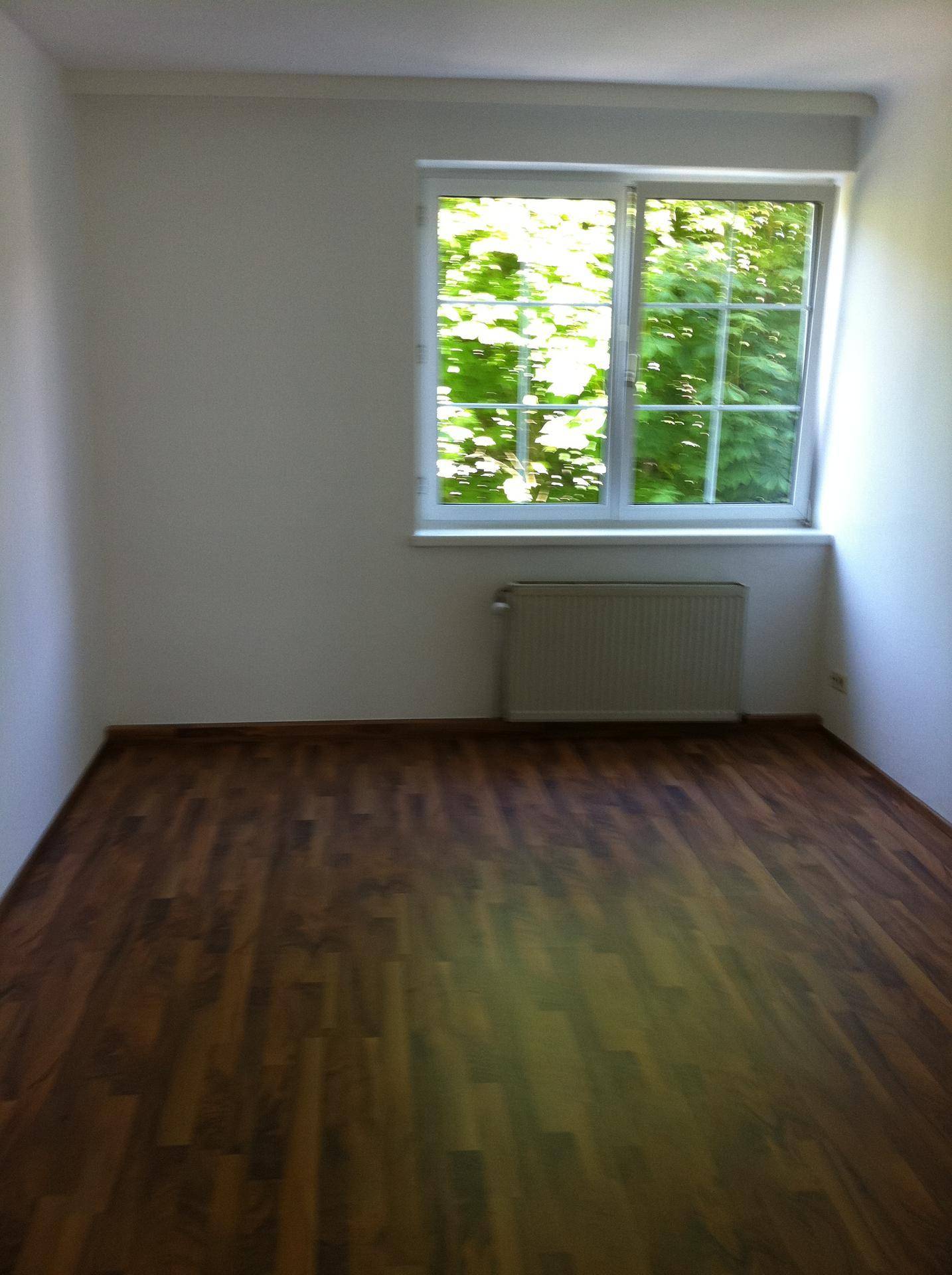 Lichtdurchfluteter, leerer Raum mit Laminatboden und einem großen Fenster mit Blick ins Grüne.