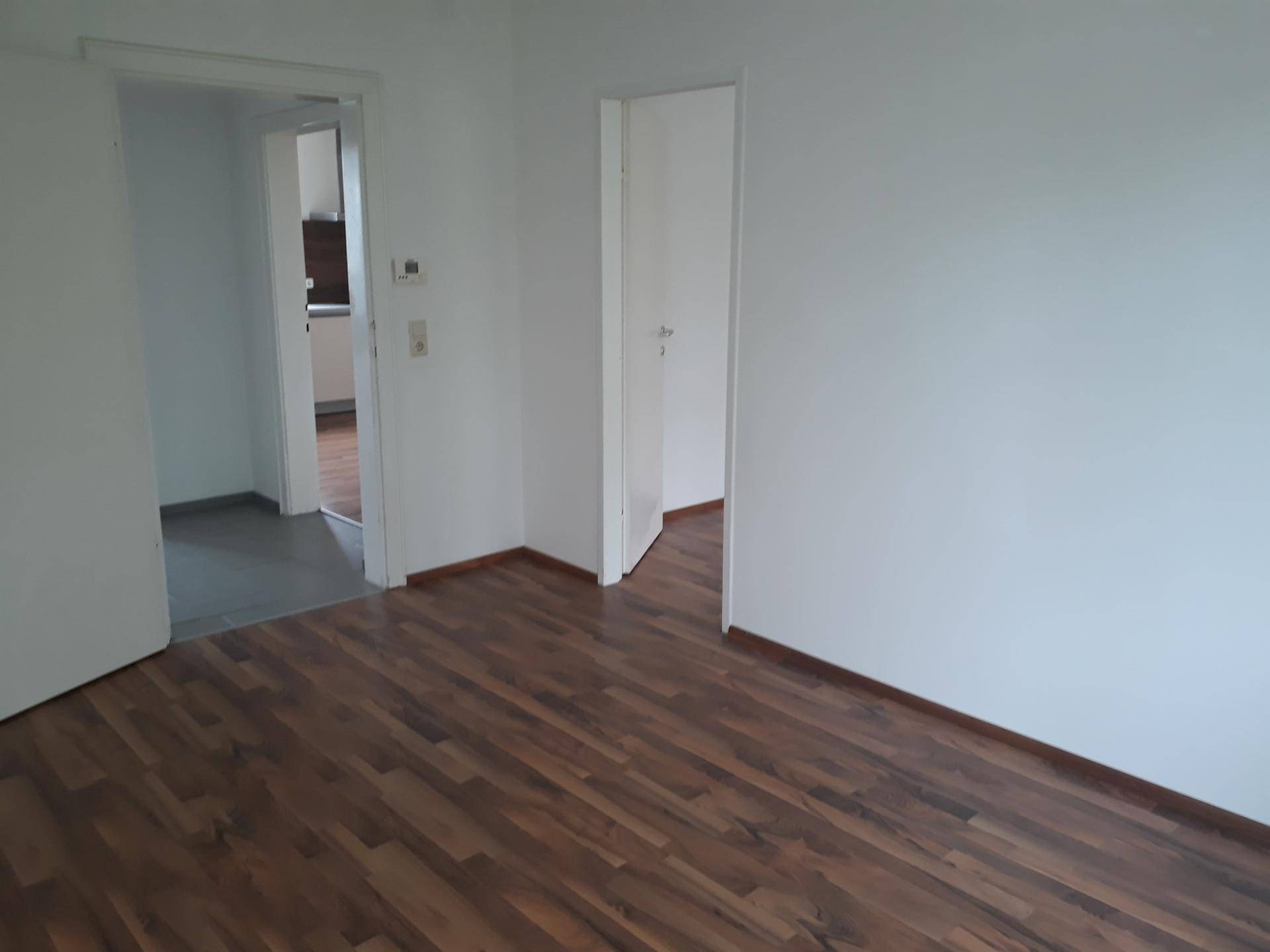 Geräumiger, leerer Raum mit Laminatboden und weißen Wänden, der Zugänge zu weiteren Bereichen bietet.