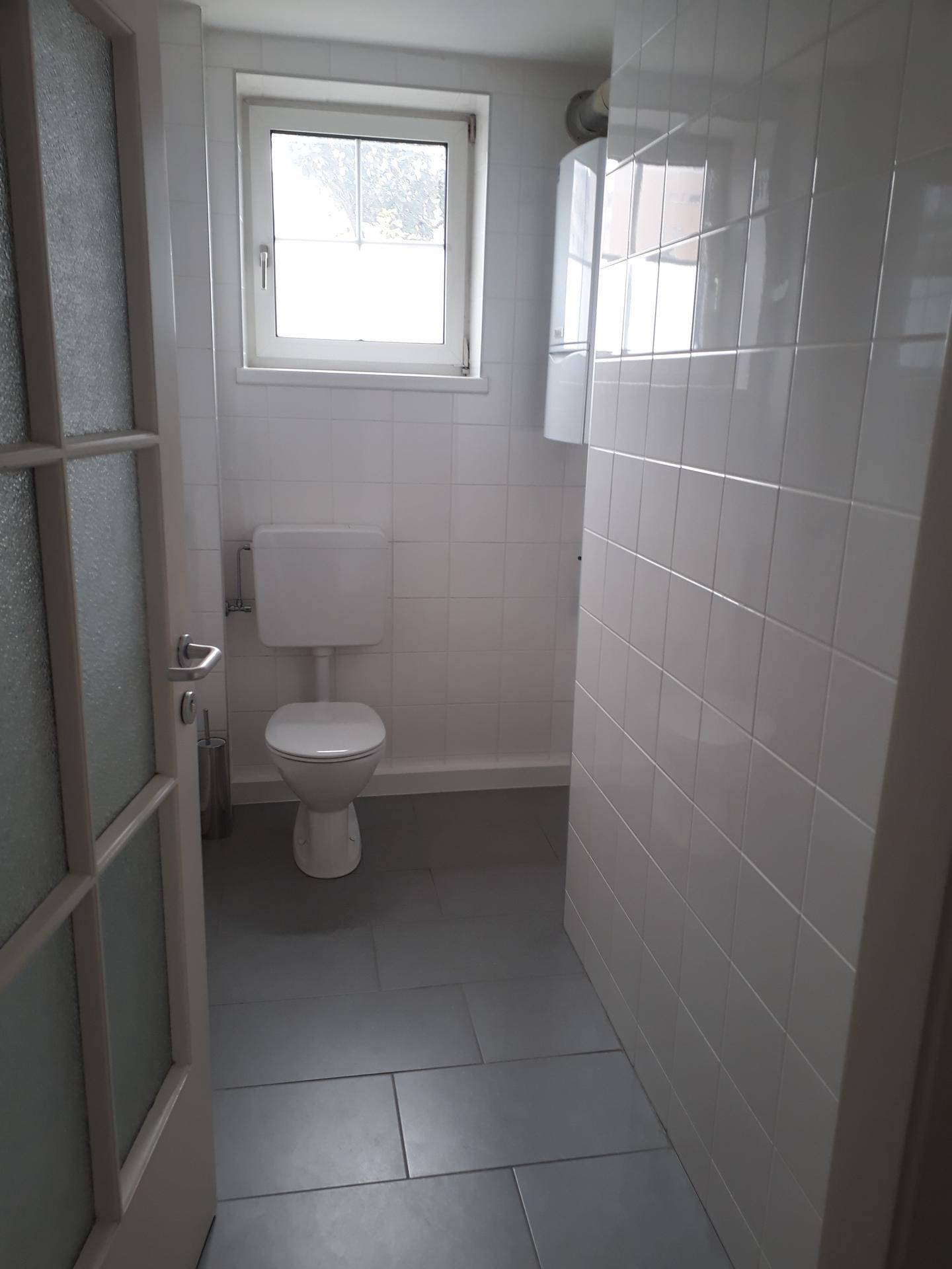 Separates WC mit weißen Wandfliesen, grauen Bodenfliesen und einem Fenster für natürliche Belüftung.