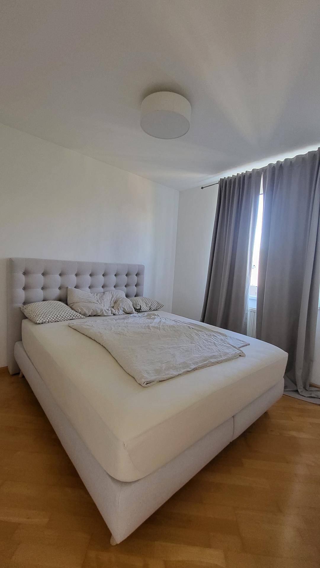 Helles Schlafzimmer mit großem Doppelbett, Parkettboden und Vorhängen.