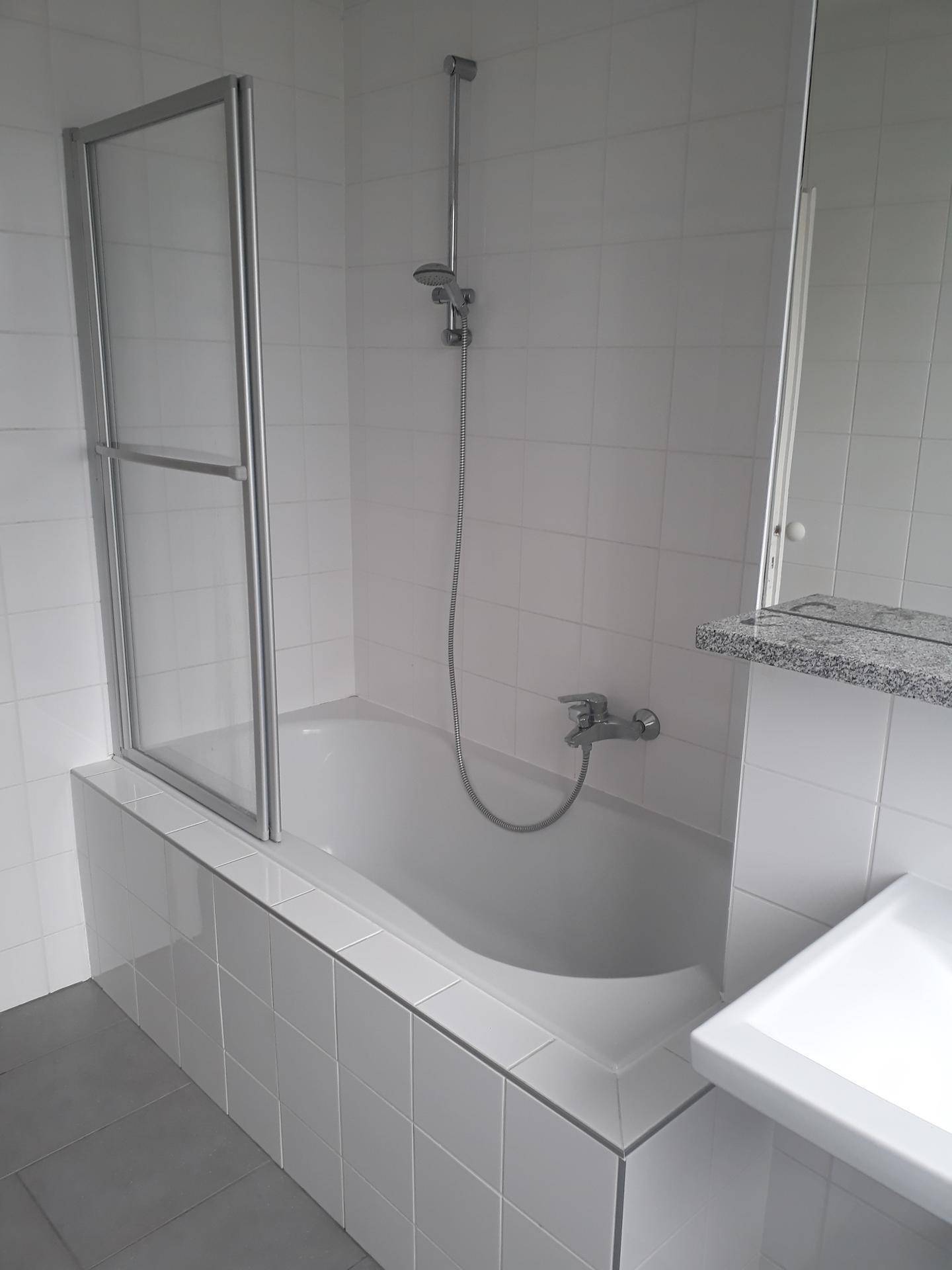 Badewanne mit Duschvorrichtung und Glasabtrennung in einem hell gefliesten Badezimmer.