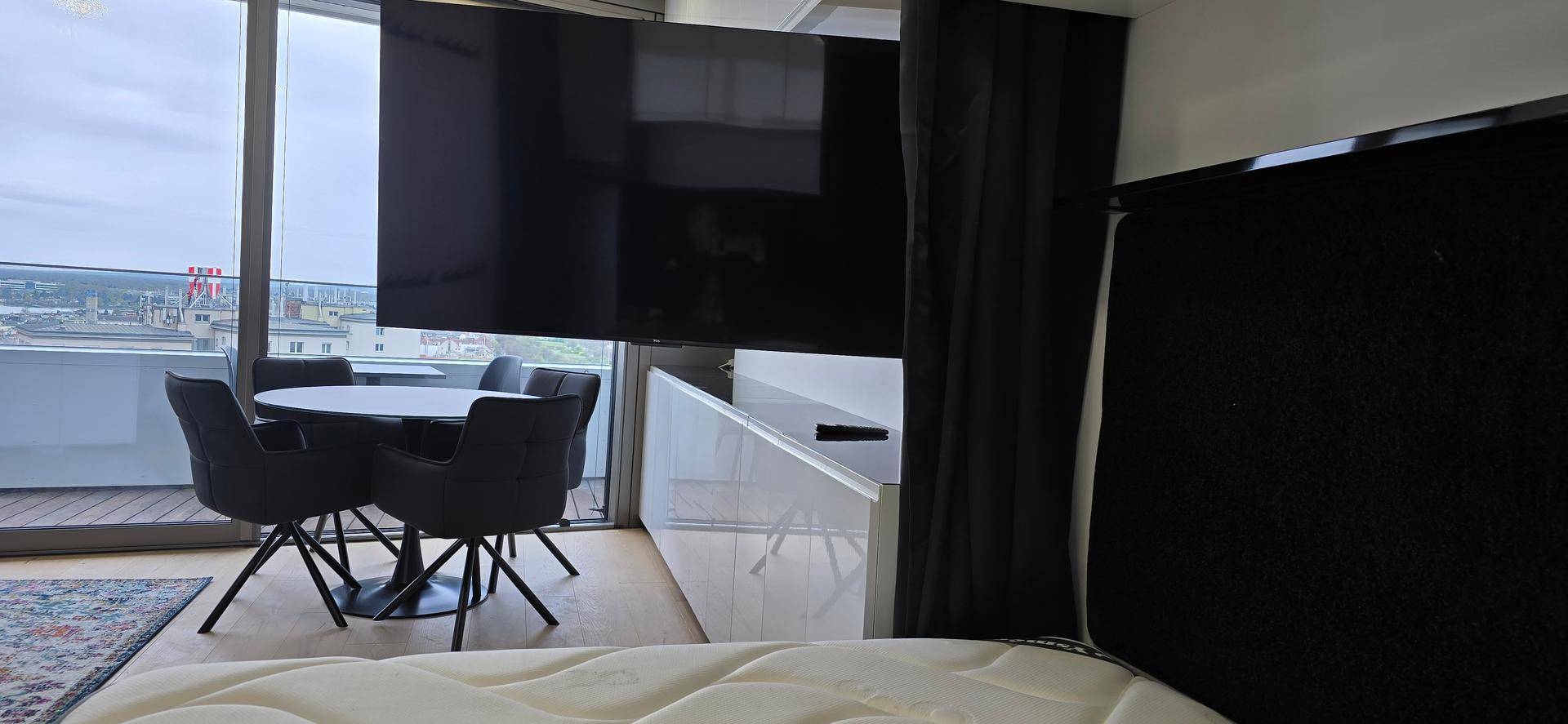 Schlaf- und Essbereich mit großem Fernseher, Esstisch und Zugang zum Balkon mit Stadtblick.