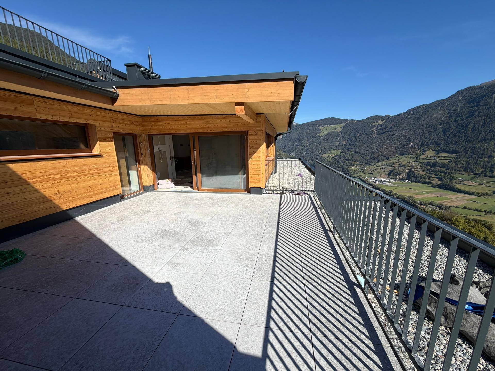 Moderne Terrasse mit Holzelementen und weitem Blick auf das Tal und die Berge unter blauem Himmel.