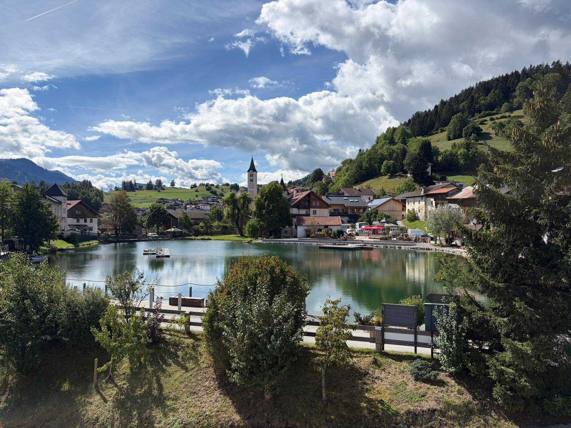 Idyllisches Dorf am See mit Kirche und grüner Berglandschaft unter bewölktem Himmel.
