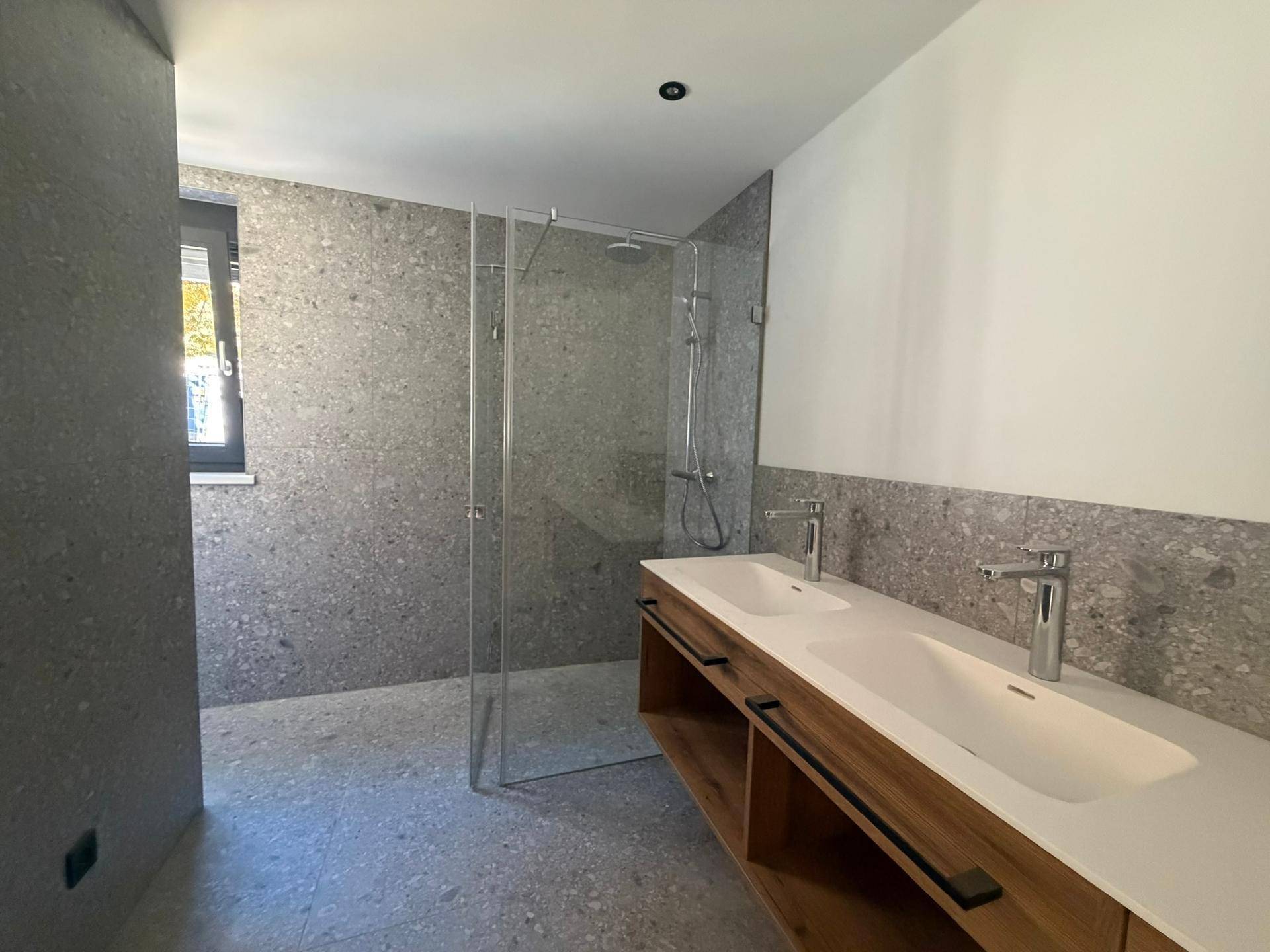 Modernes Badezimmer mit grauen Fliesen, begehbarer Dusche und Doppelwaschbecken mit Holzunterschrank.