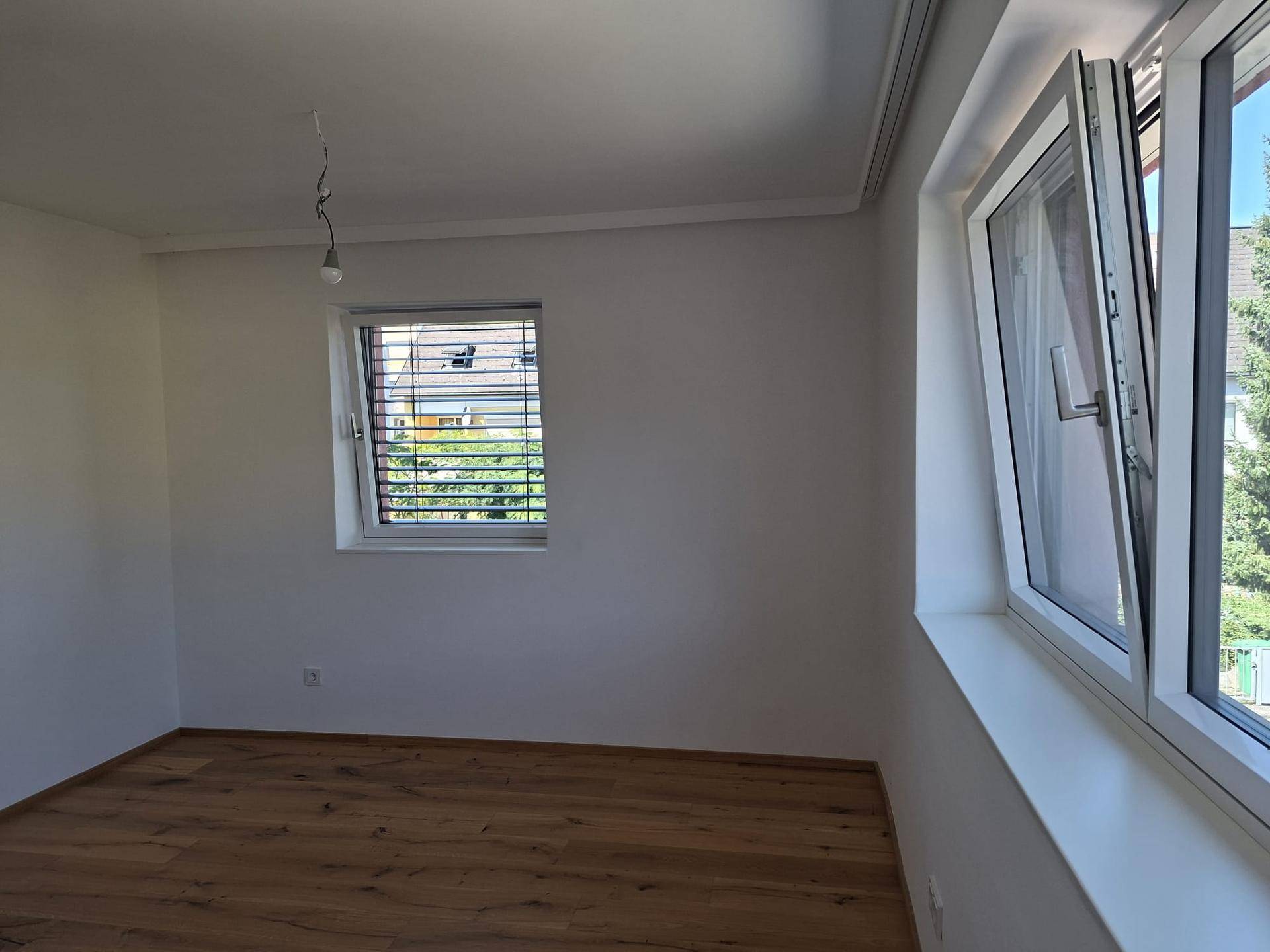 Helles Zimmer mit Holzboden und zwei Fenstern, die viel Tageslicht hereinlassen.