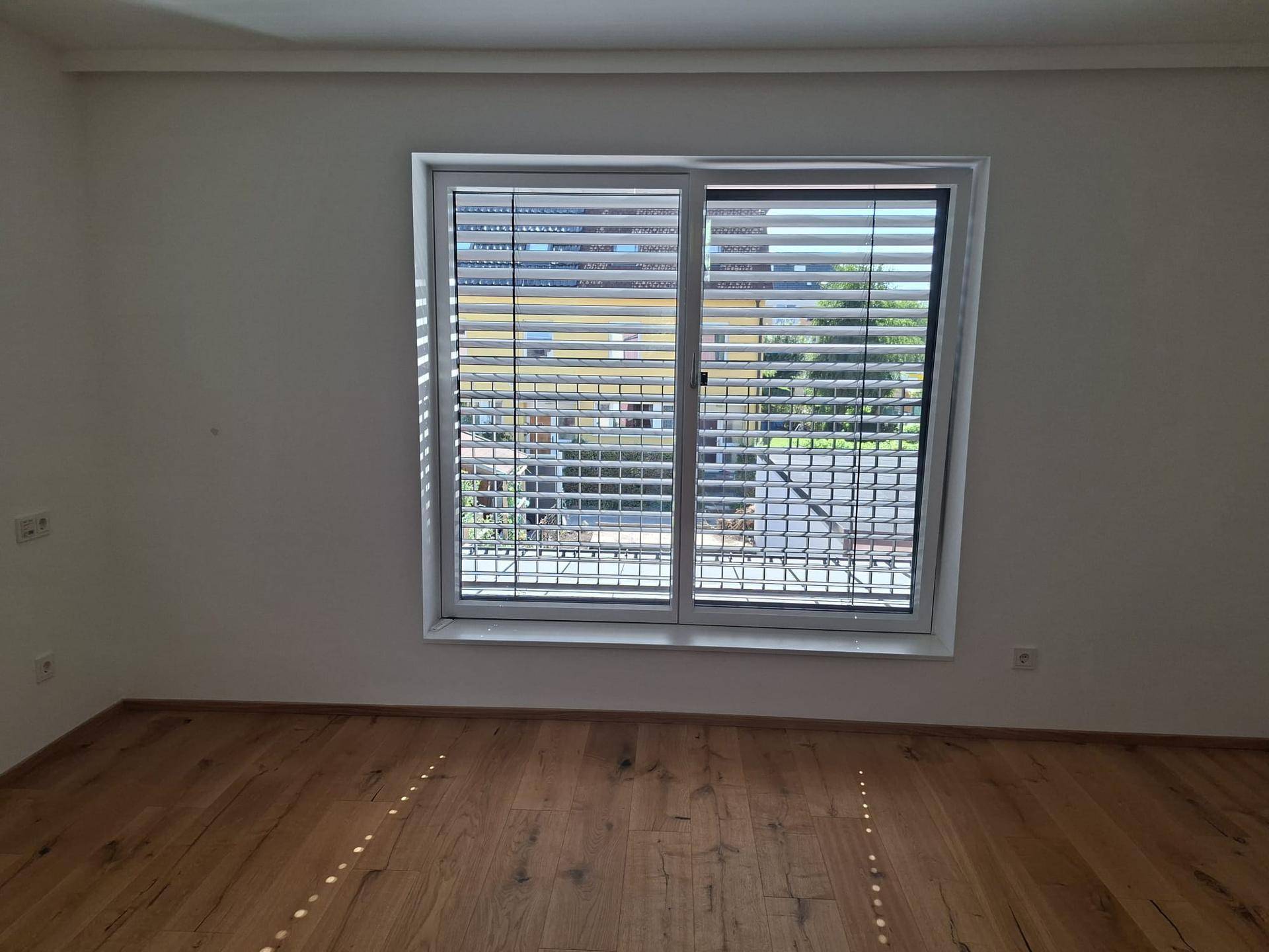 Helles Schlafzimmer mit Holzfußboden und großem Fenster mit Jalousien.