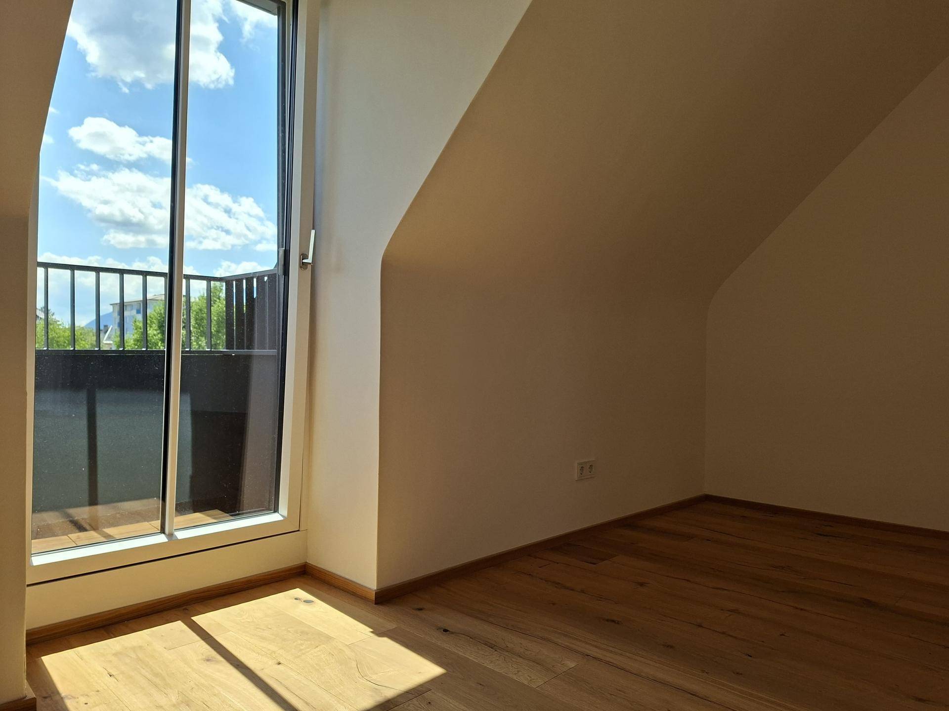 Helles Dachzimmer mit Holzboden, schrägen Wänden und Zugang zu einem Balkon.