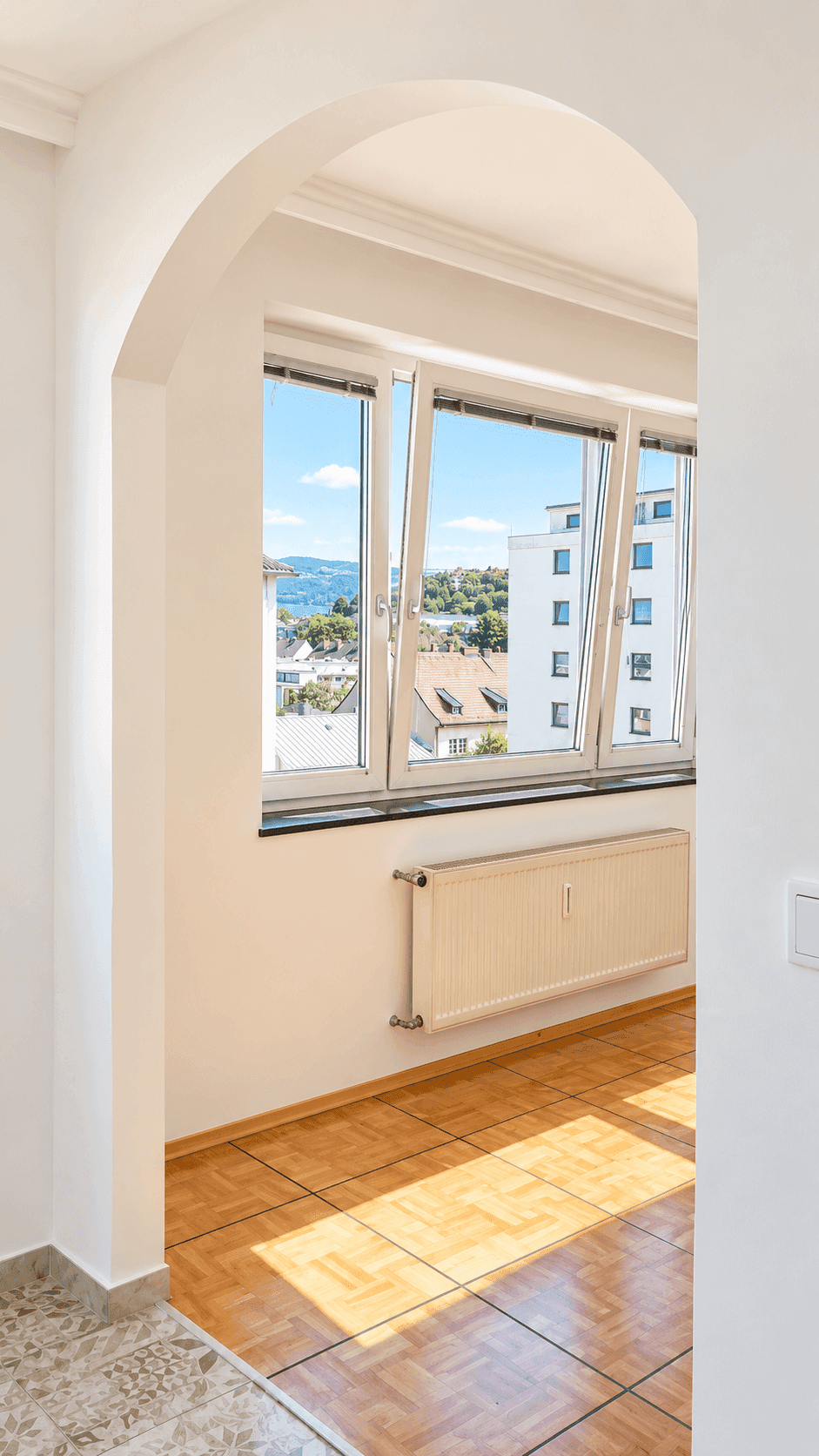 Heller Bereich mit Bogenöffnung, Parkettboden und großen Fenstern mit Blick auf die Stadt.