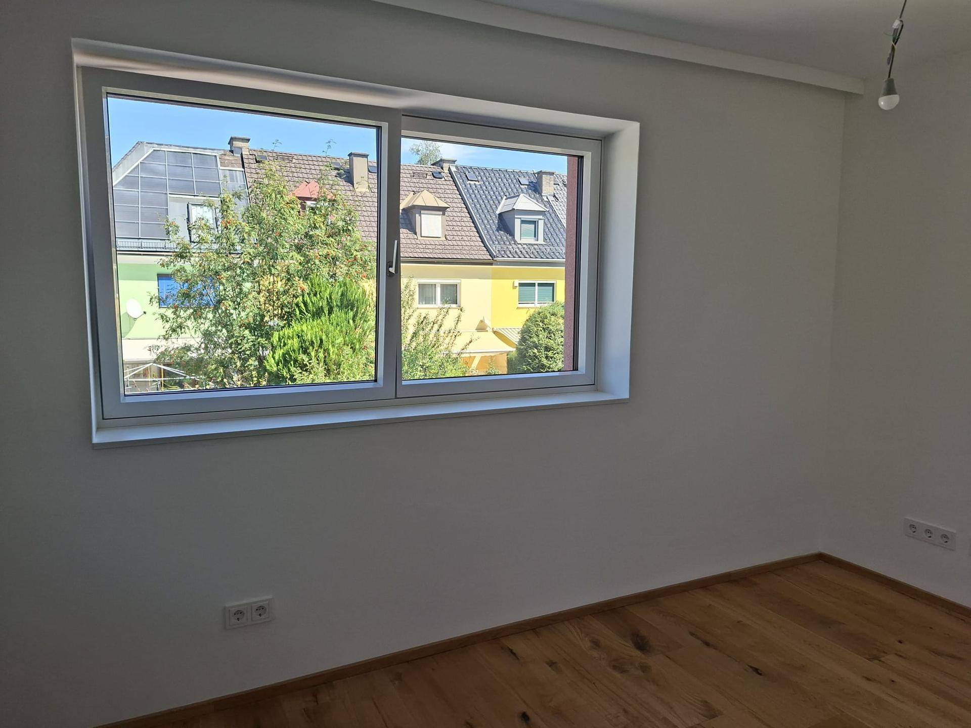 Schlafzimmer mit Holzboden und Fenster, das einen Blick auf die Nachbarhäuser bietet.