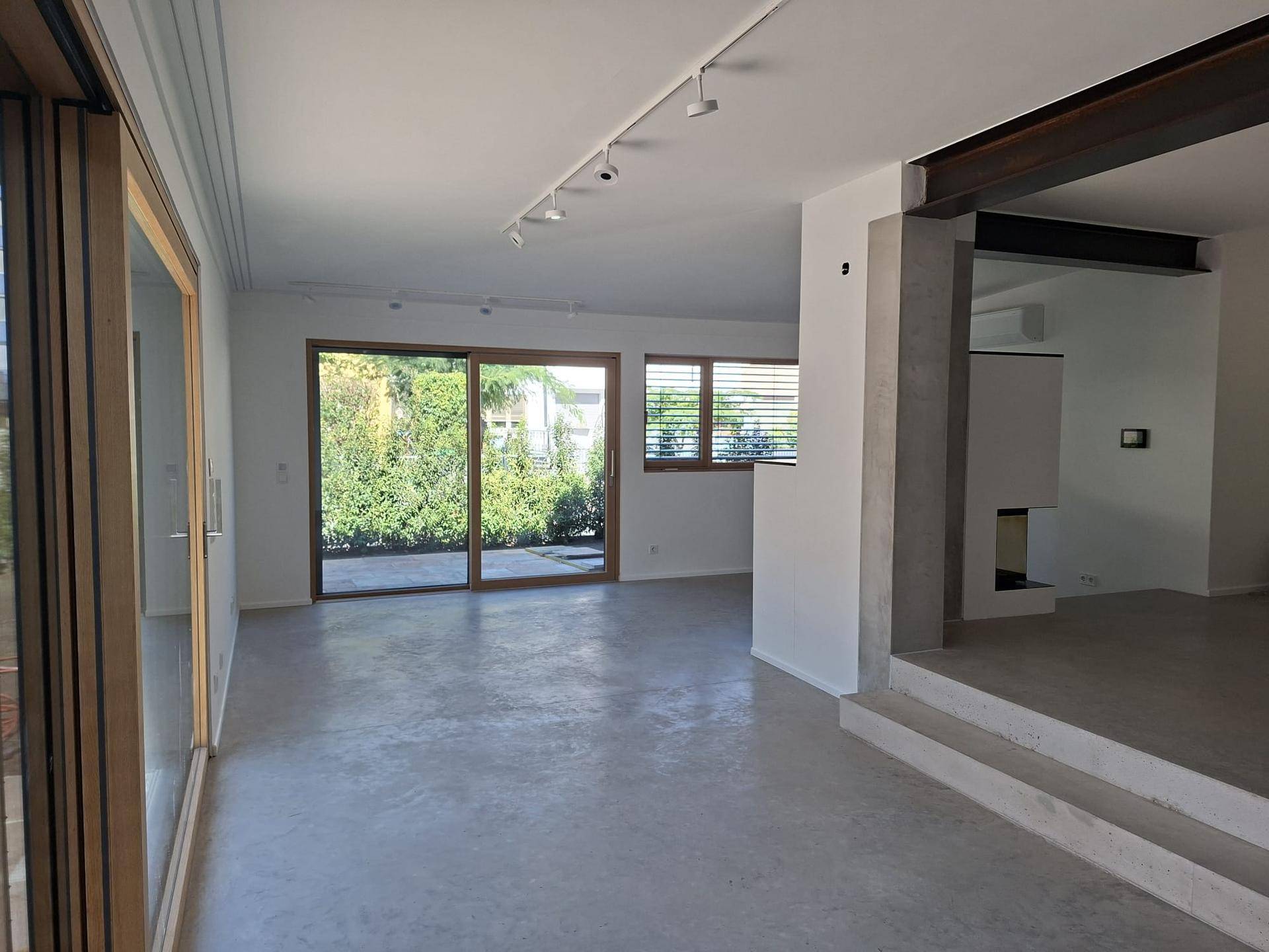 Geräumiges Wohnzimmer mit großen Fenstern, Betonboden und Blick ins Grüne.