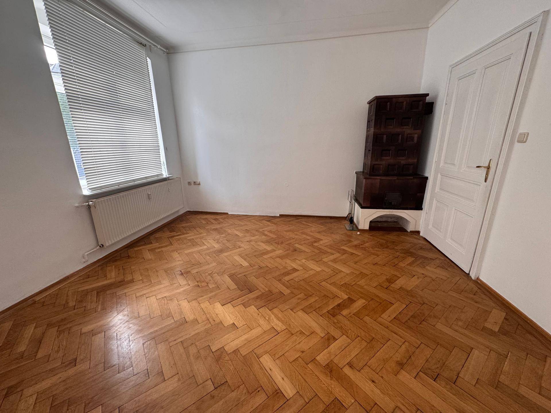 Heller Raum mit Fischgrätparkett und einem traditionellen Kachelofen, ideal als Wohn- oder Schlafzimmer.