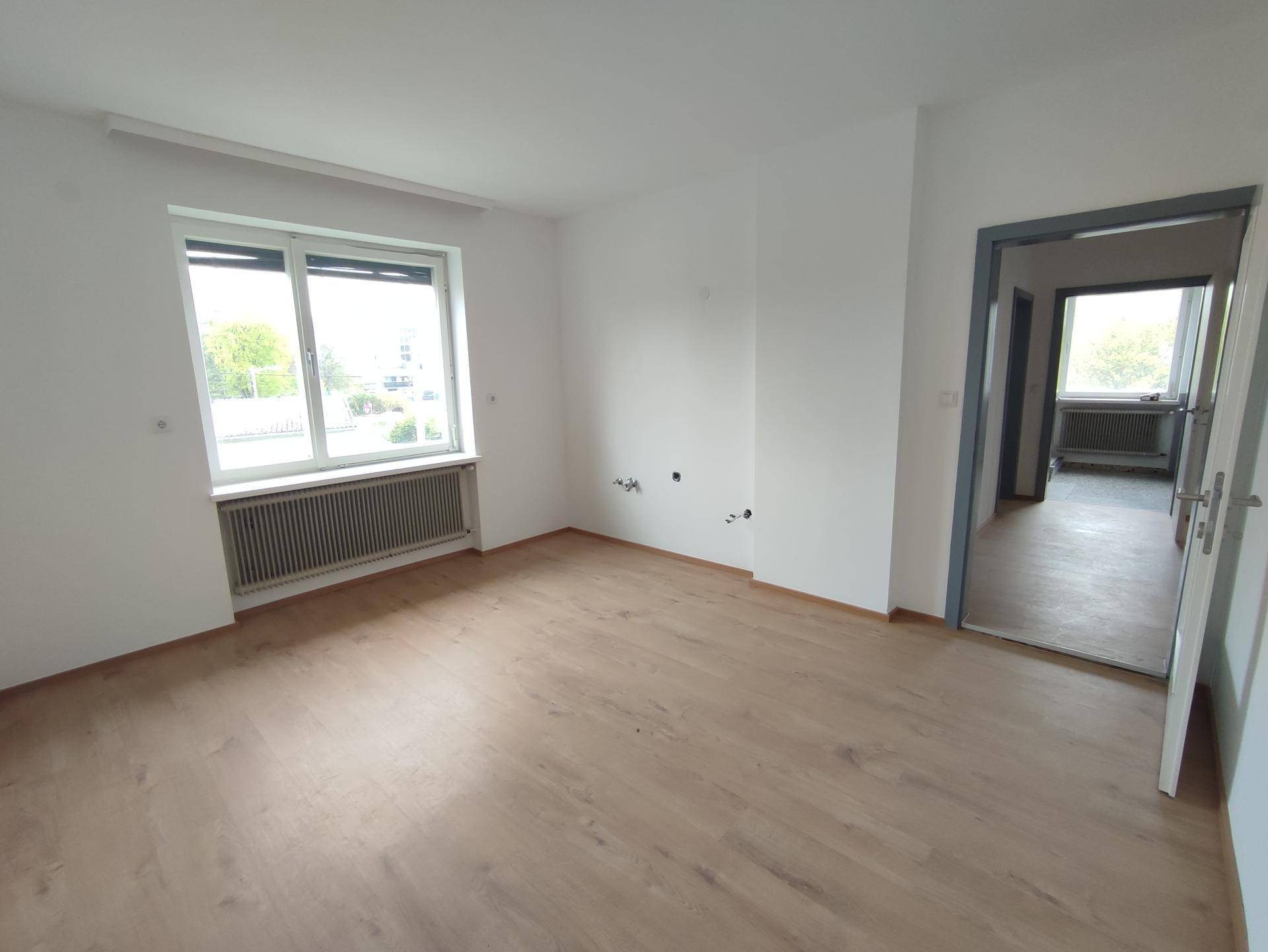 Heller Wohnraum mit Laminatboden, großem Fenster und Zugang zu einem weiteren Zimmer.