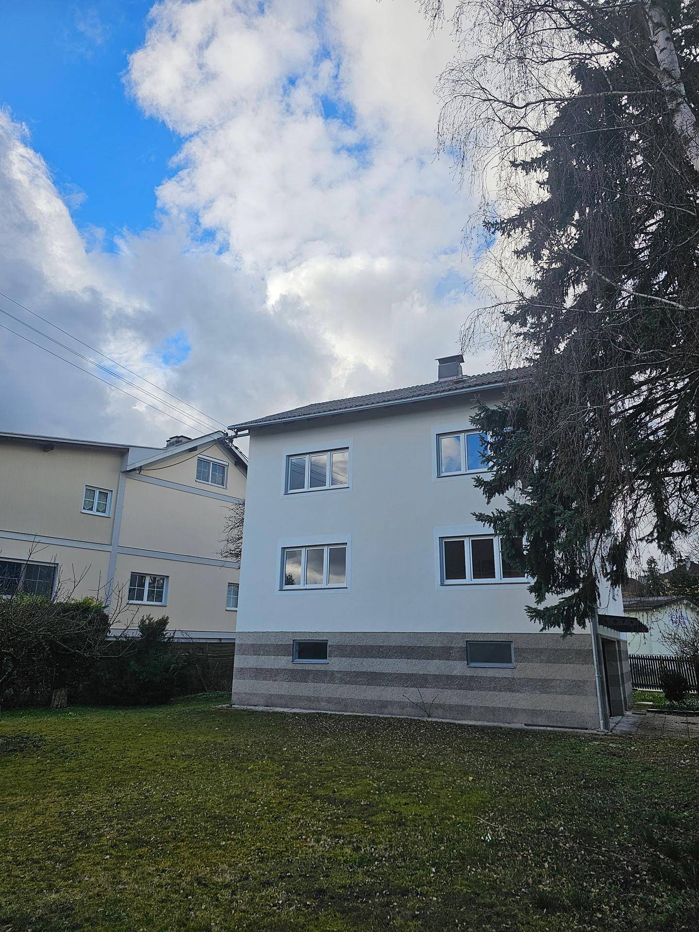 Seitenansicht des renovierten Gebäudes mit grüner Umgebung und hohen Bäumen unter blauem Himmel.