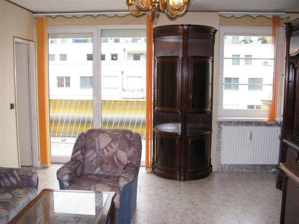 Wohnzimmer mit Fliesenboden, gemustertem Sessel und großem Holzschrank, bietet Zugang zum Balkon.