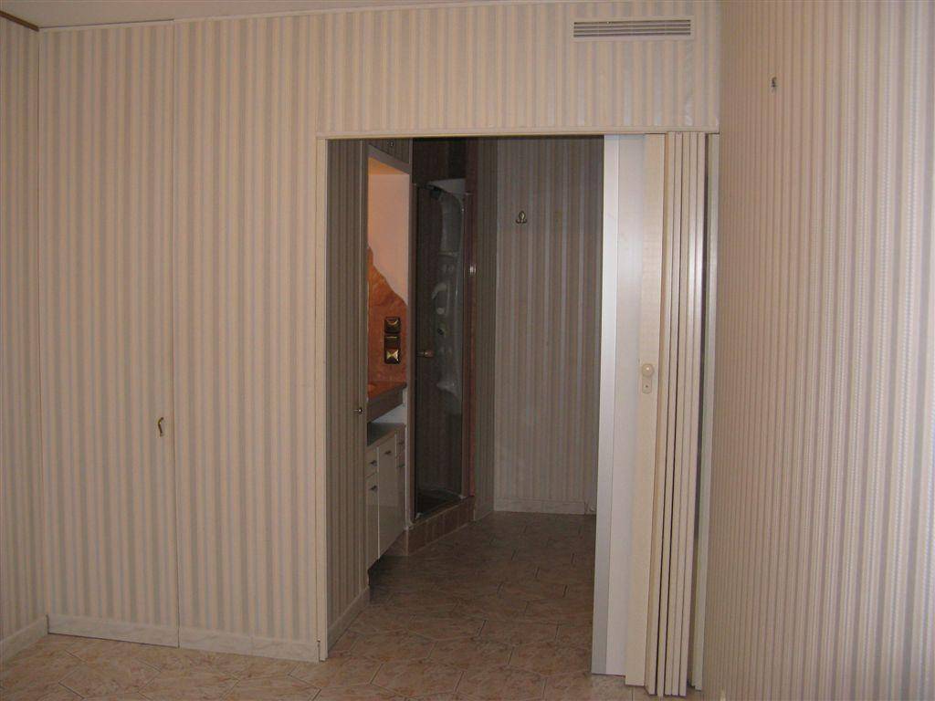 Flur mit gestreifter Tapete und Falttür, führt zu einem Badezimmer mit Duschbereich und Waschbecken.