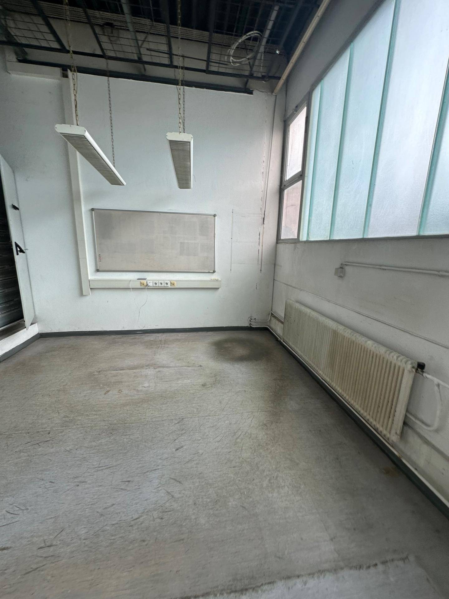Leerer Raum mit Betonboden, Heizkörper und großen Fenstern, die viel Tageslicht hereinlassen.