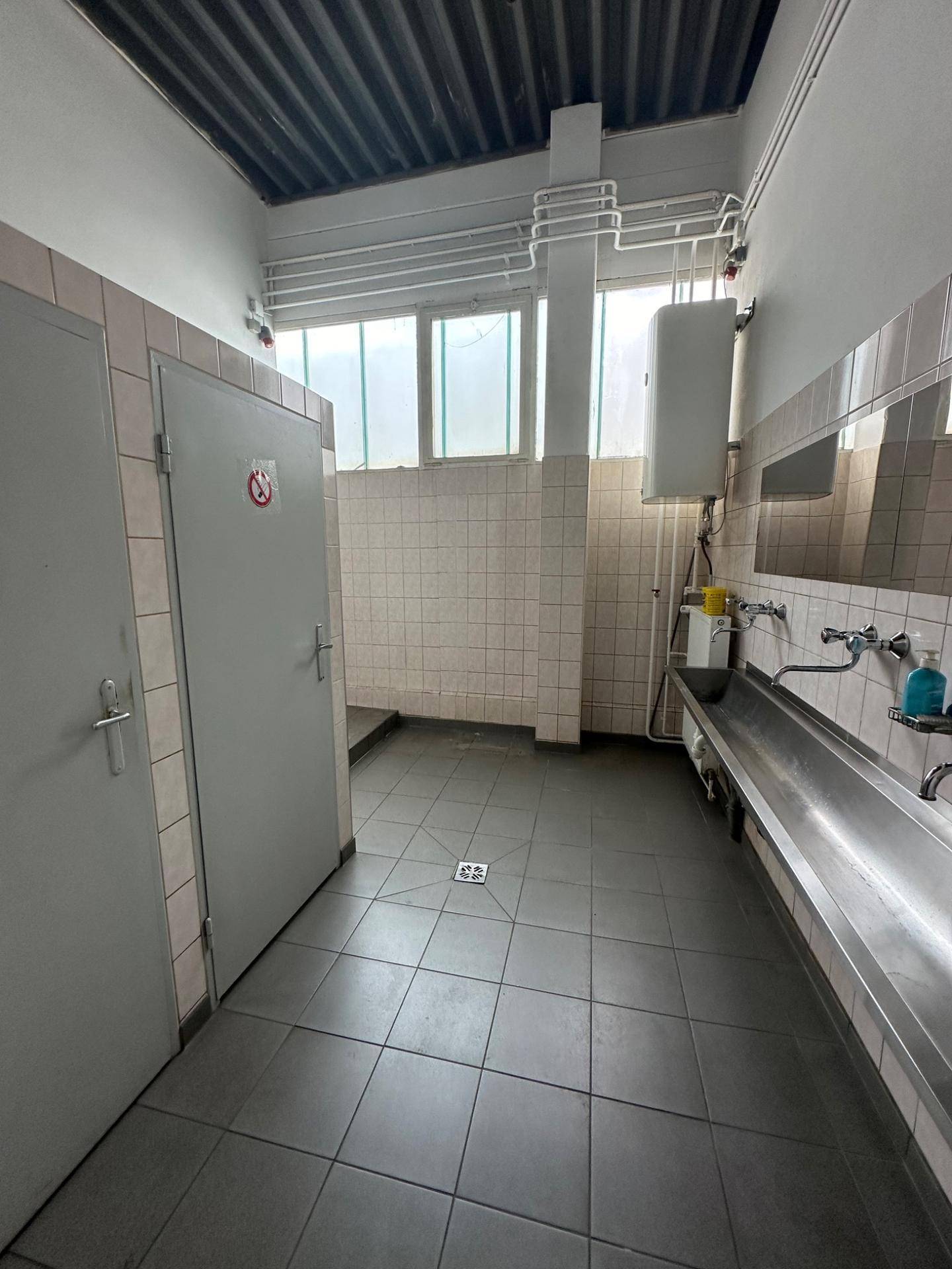 Industrieller Waschraum mit gefliestem Boden, mehreren Toilettenkabinen und einem langen Waschbecken.