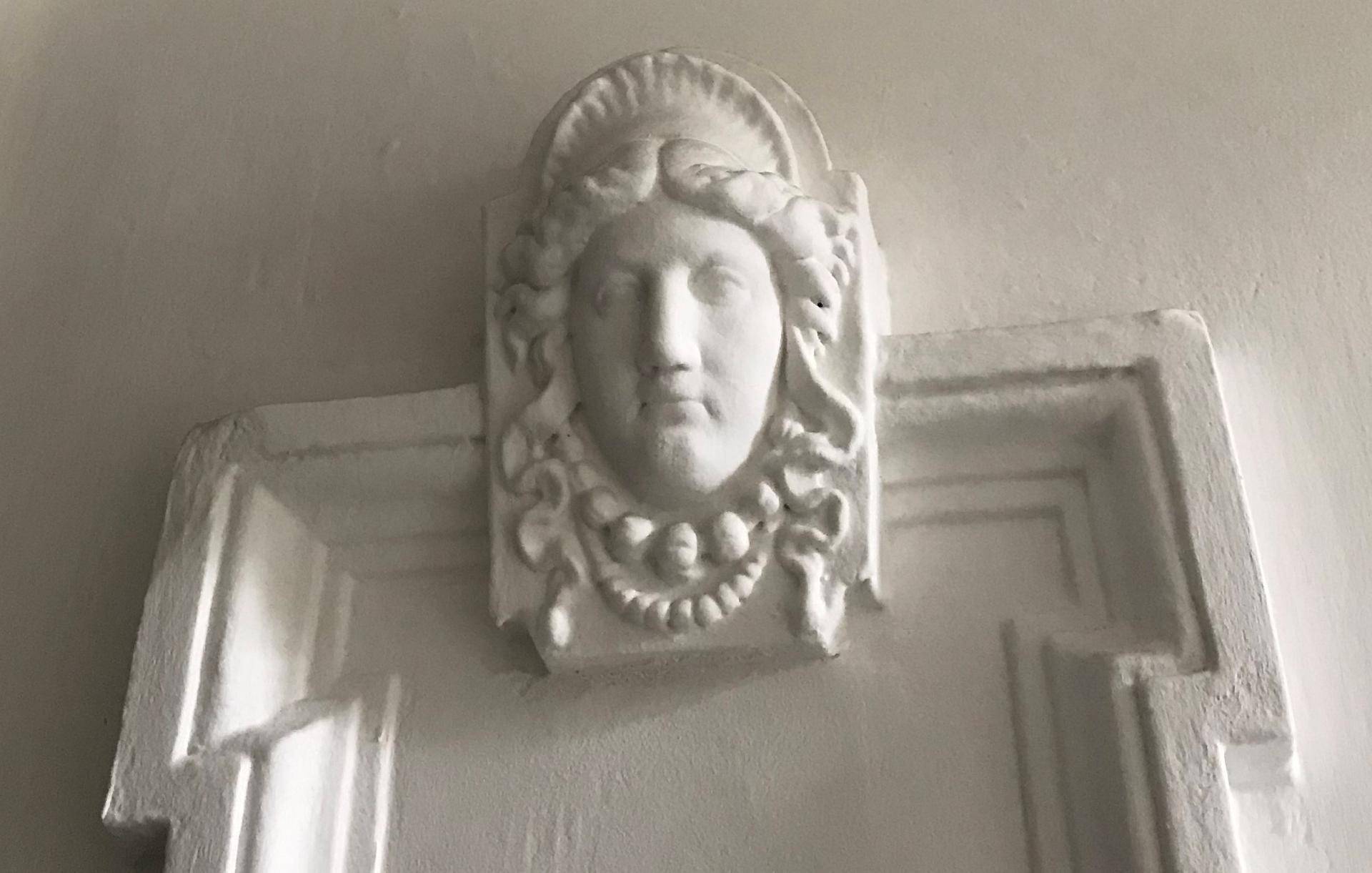 Detailaufnahme eines weißen Stuckreliefs mit klassischem Gesichtsmotiv an der Wand.