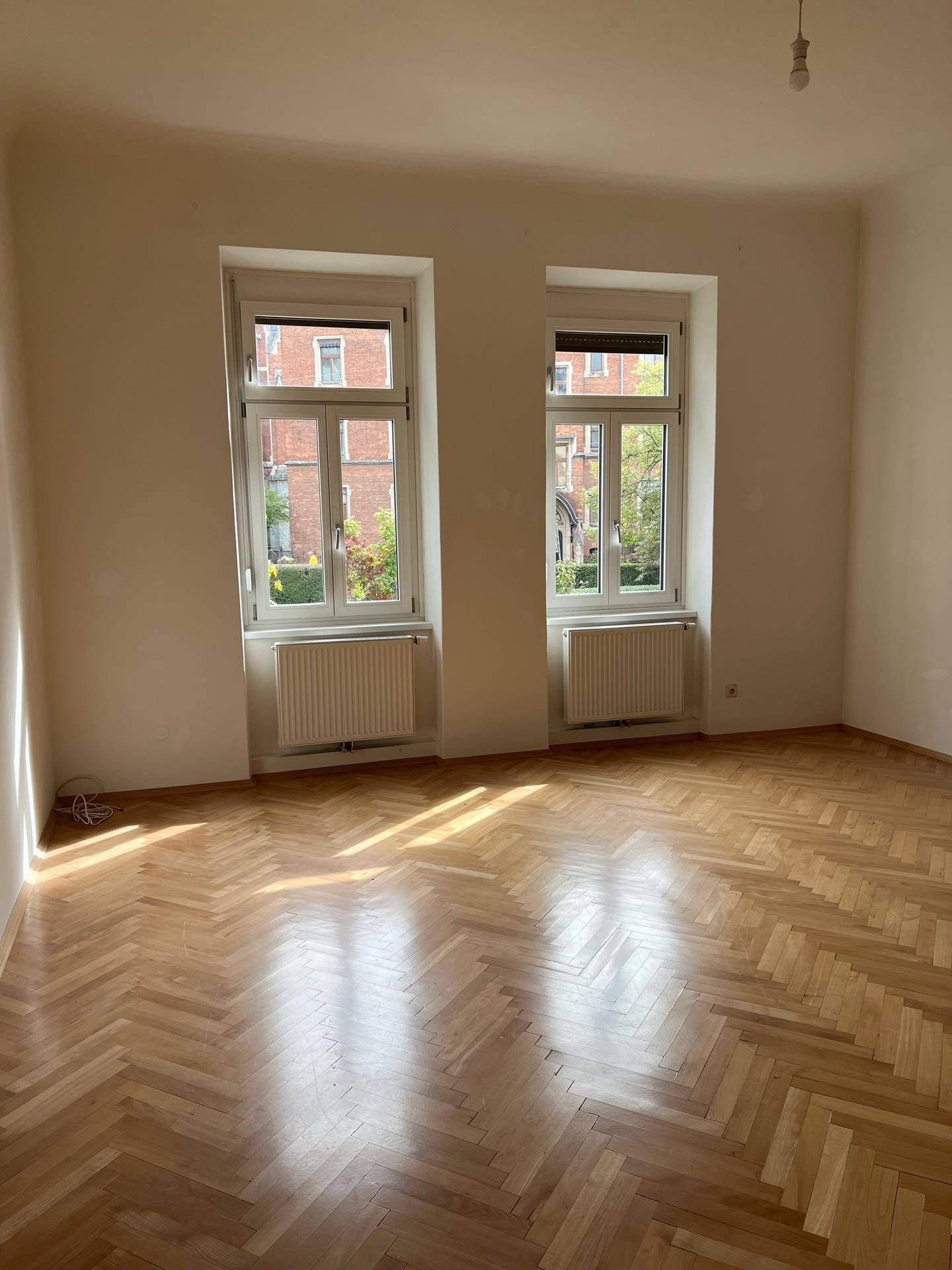 Helles, leeres Zimmer mit Fischgrätparkettboden, zwei Fenstern und viel natürlichem Licht.