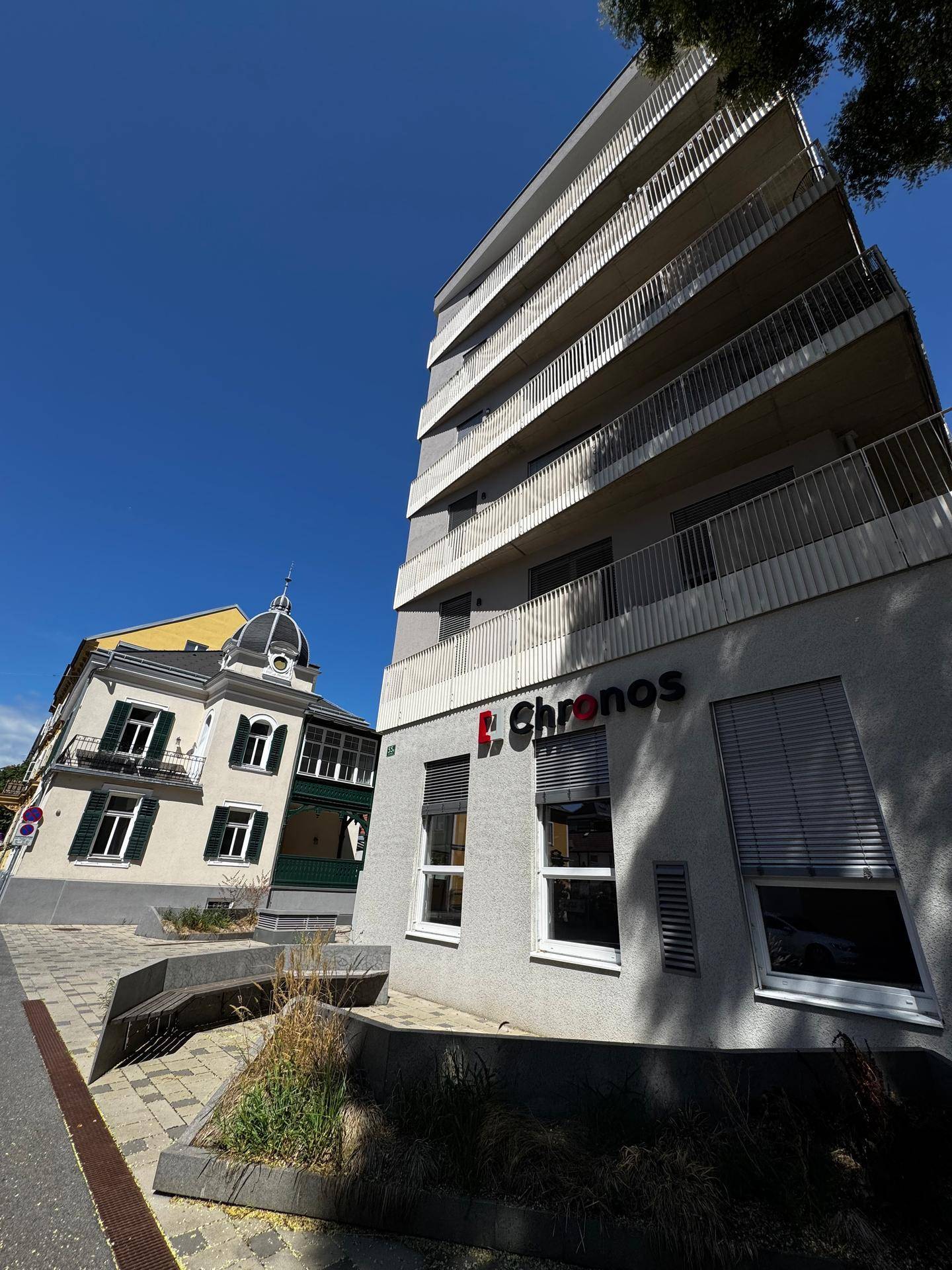 Modernes Wohngebäude mit Balkonen und angrenzendem Altbau unter strahlend blauem Himmel.