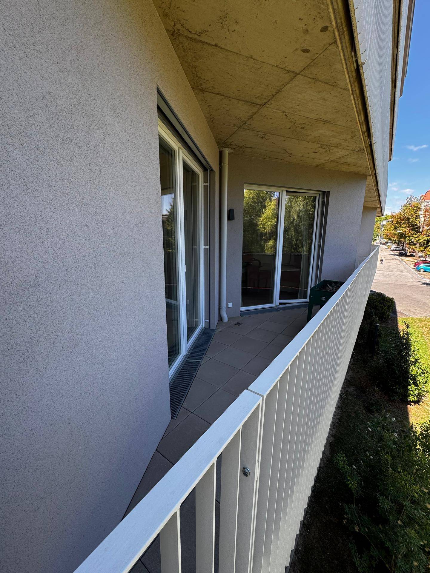 Langer Balkon mit Fliesenboden und Blick auf die Straße sowie umliegende Bäume.