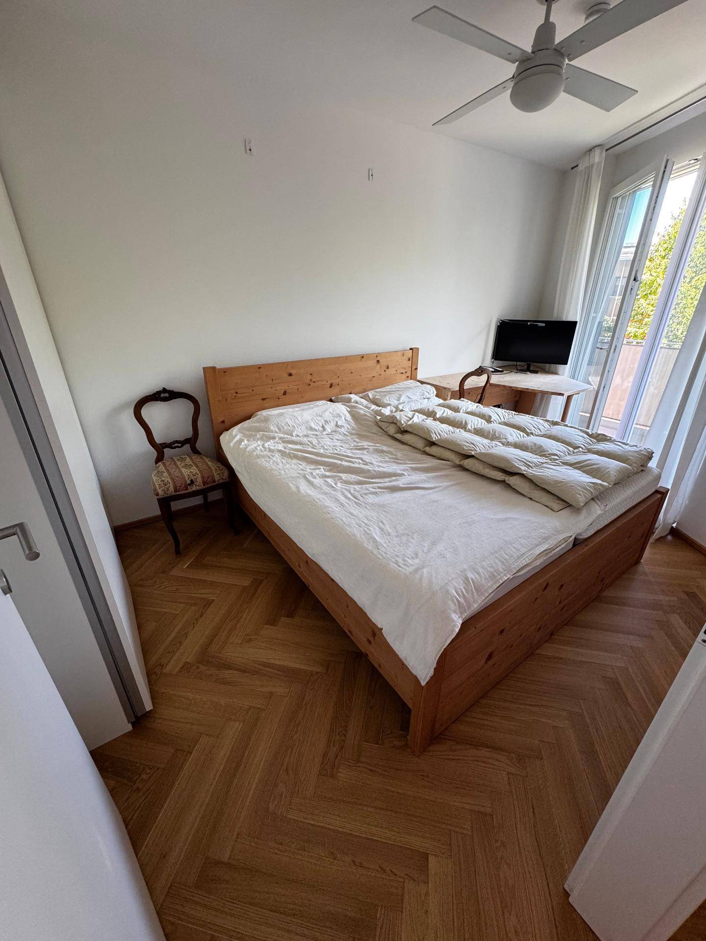 Helles Schlafzimmer mit Holzbett, Deckenventilator und Zugang zu einem sonnigen Balkon.