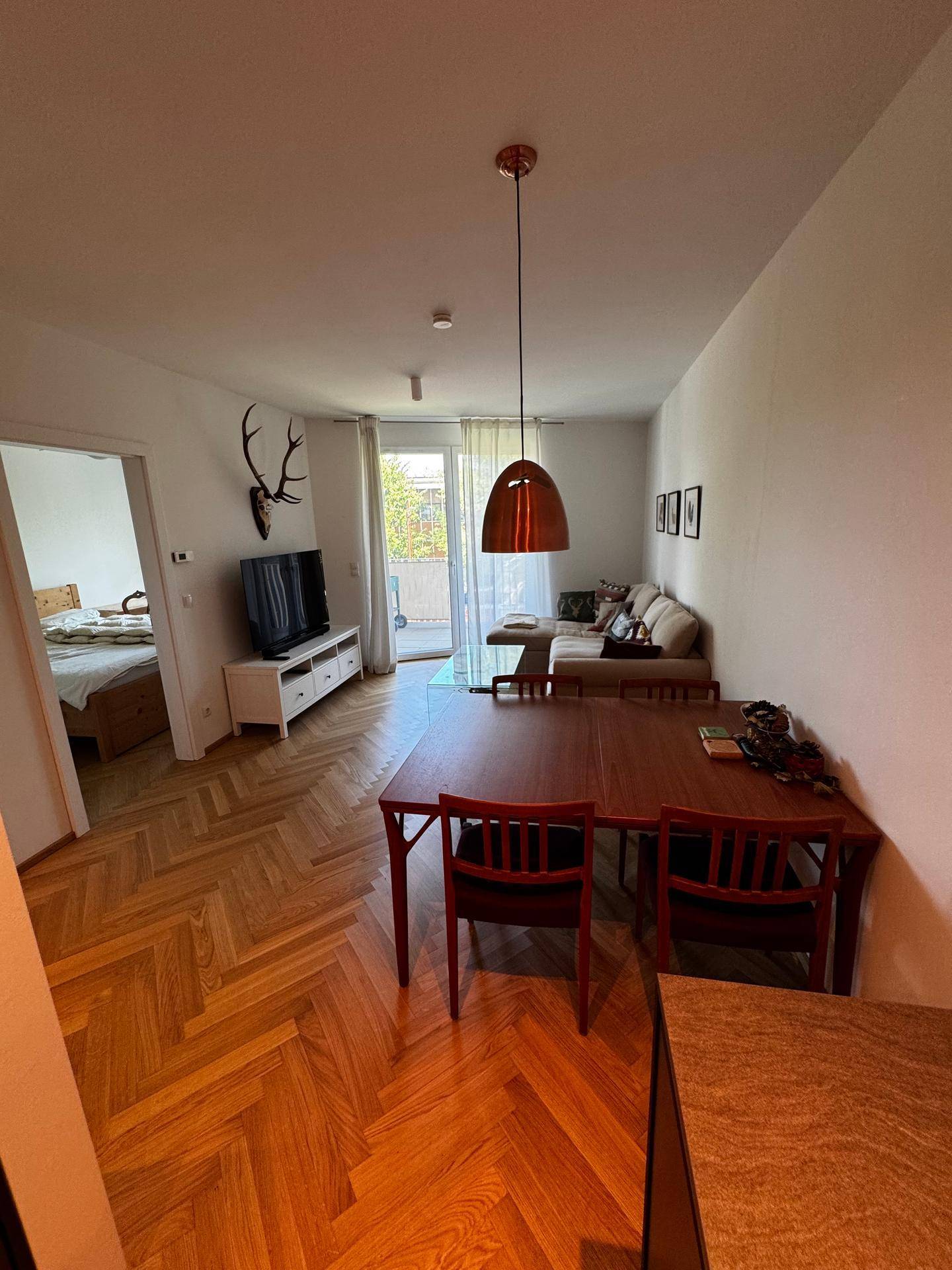 Geräumiger Wohn- und Essbereich mit Fischgrätparkett, Esstisch und Zugang zum Schlafzimmer.
