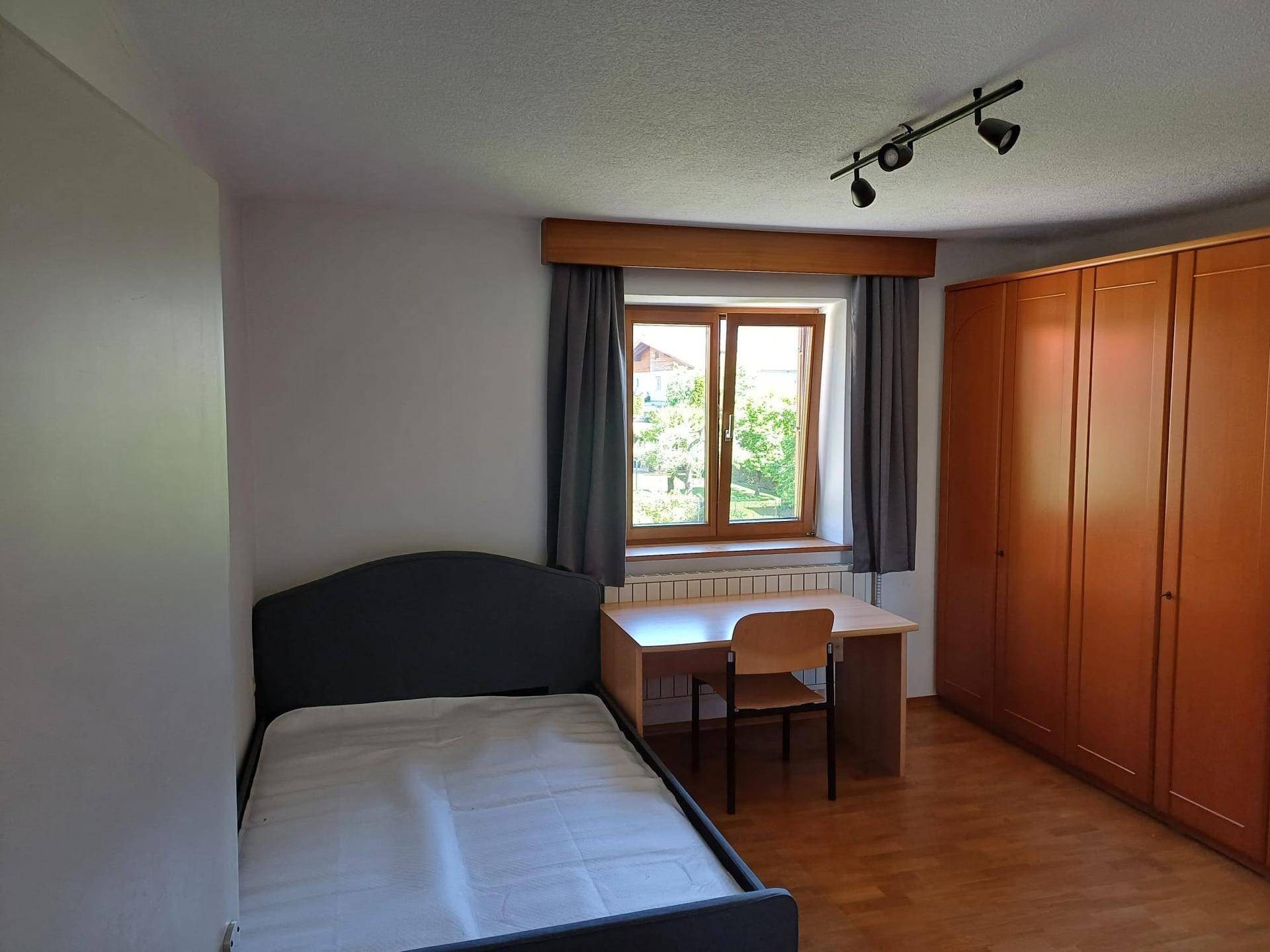 Schlafzimmer mit Einzelbett, Schreibtisch und großem Kleiderschrank, hell durch ein Fenster.