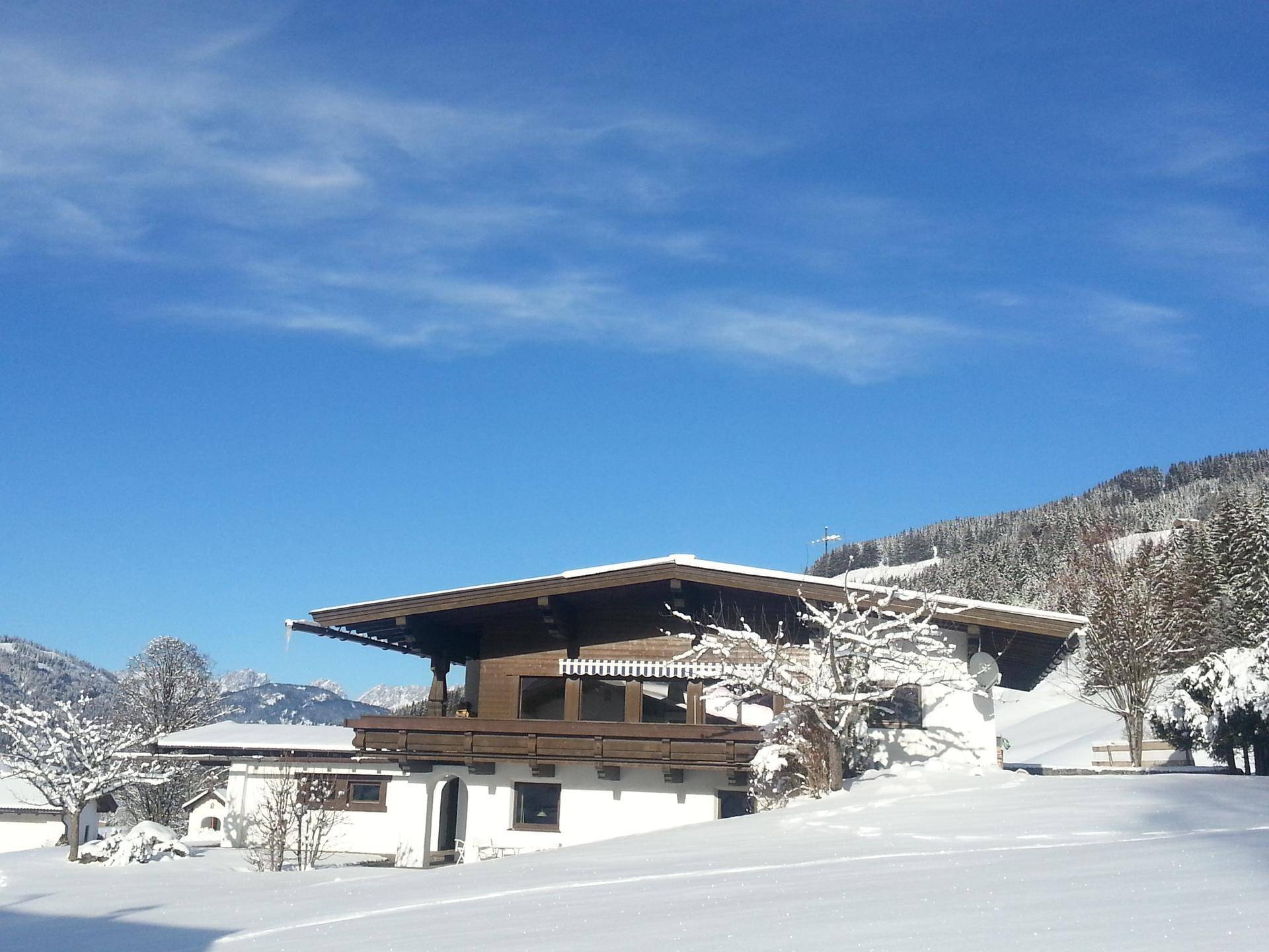 Das Haus im Winter, umgeben von Schnee, mit traditionellen Holzelementen und weißer Fassade unter blauem Himmel.