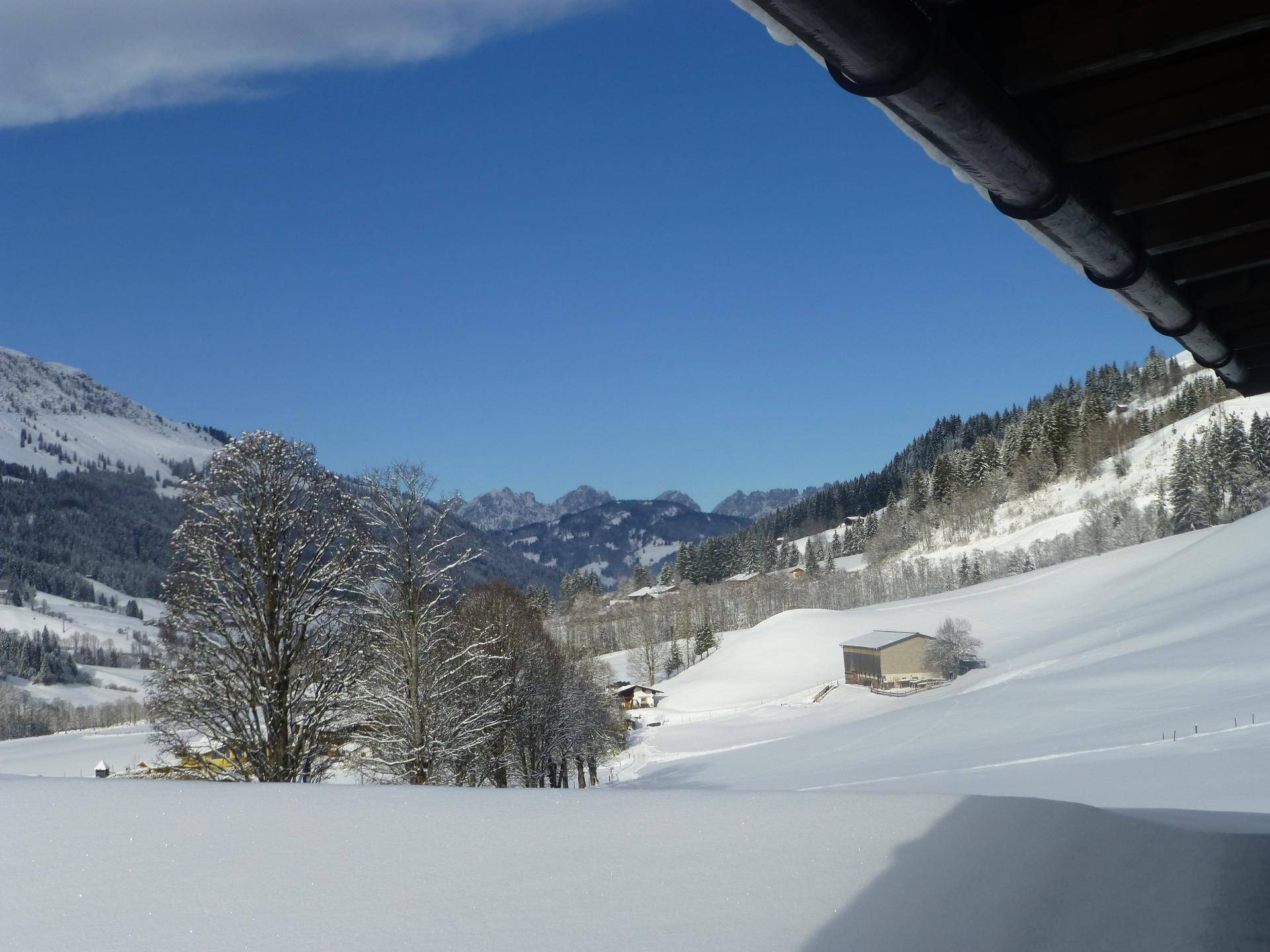 Malerische Winterlandschaft mit tief verschneiten Hügeln, Bäumen und einer Hütte vor einer Bergkulisse.
